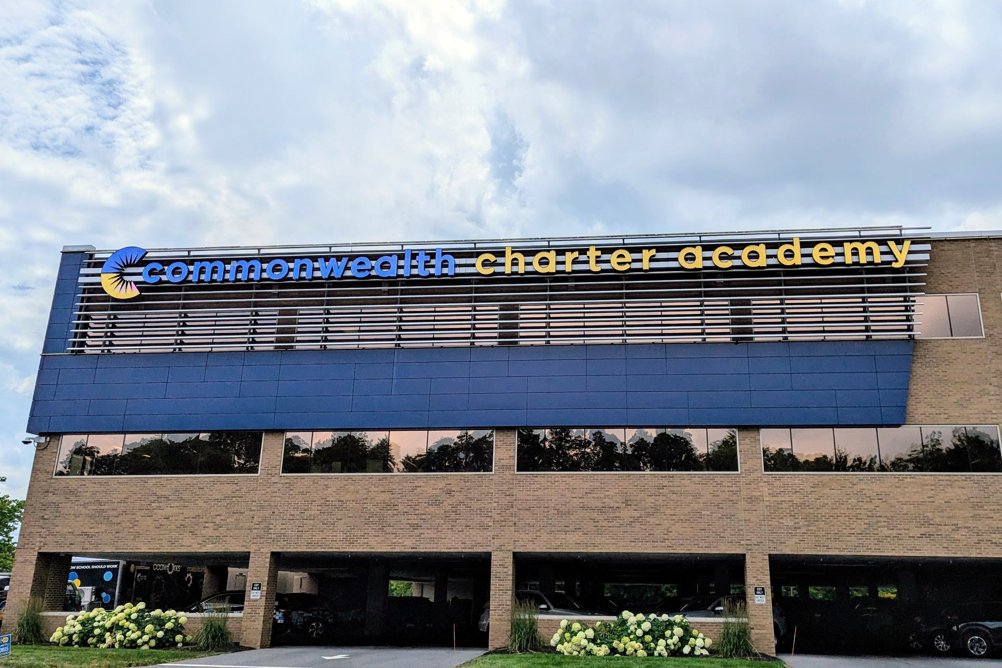 A photograph of a large tan and blue stone building with the words "Commonwealth Charter Academy" across the top and a cloudy sky in the background.