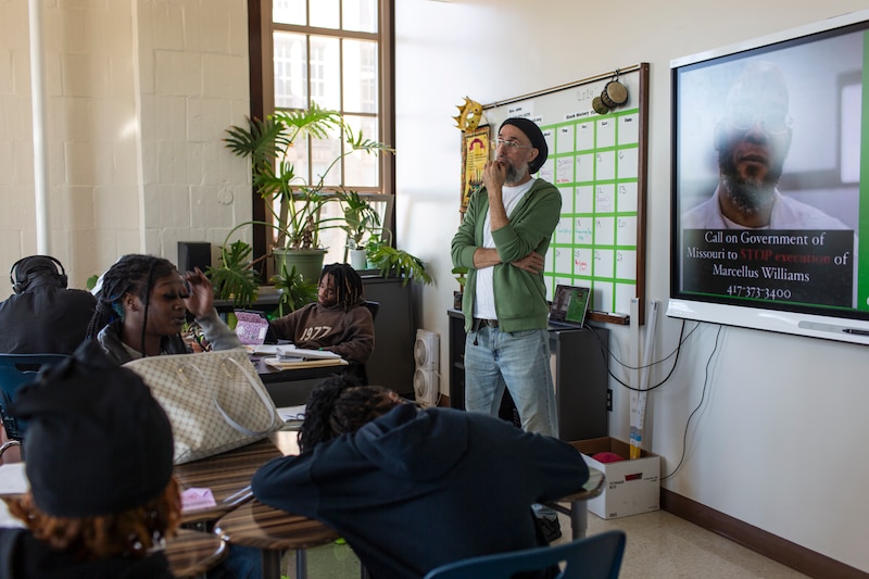 A teacher wearing a green sweater teaches next to a dry erase board in front of a class full of students.