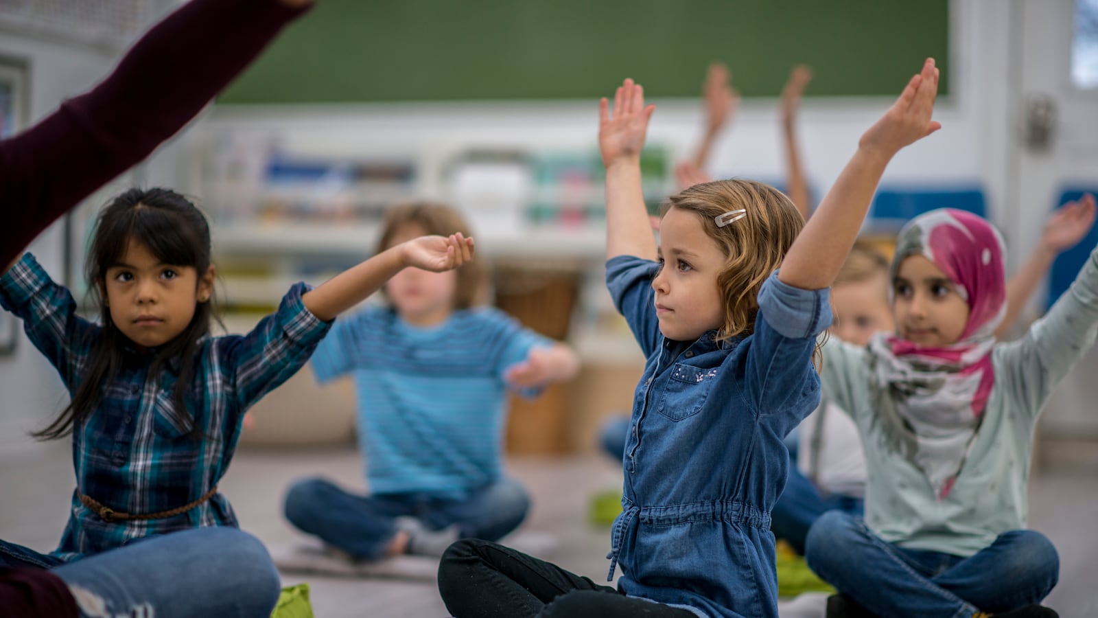 A multi-ethnic group of young school children are indoors in their classroom. They are sitting on pillows and doing yoga together. They are stretching their arms out above their heads.