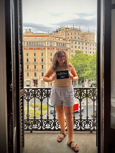 A photograph of a woman holding a small black board posing for a photograph on a balcony with buildings in the background.