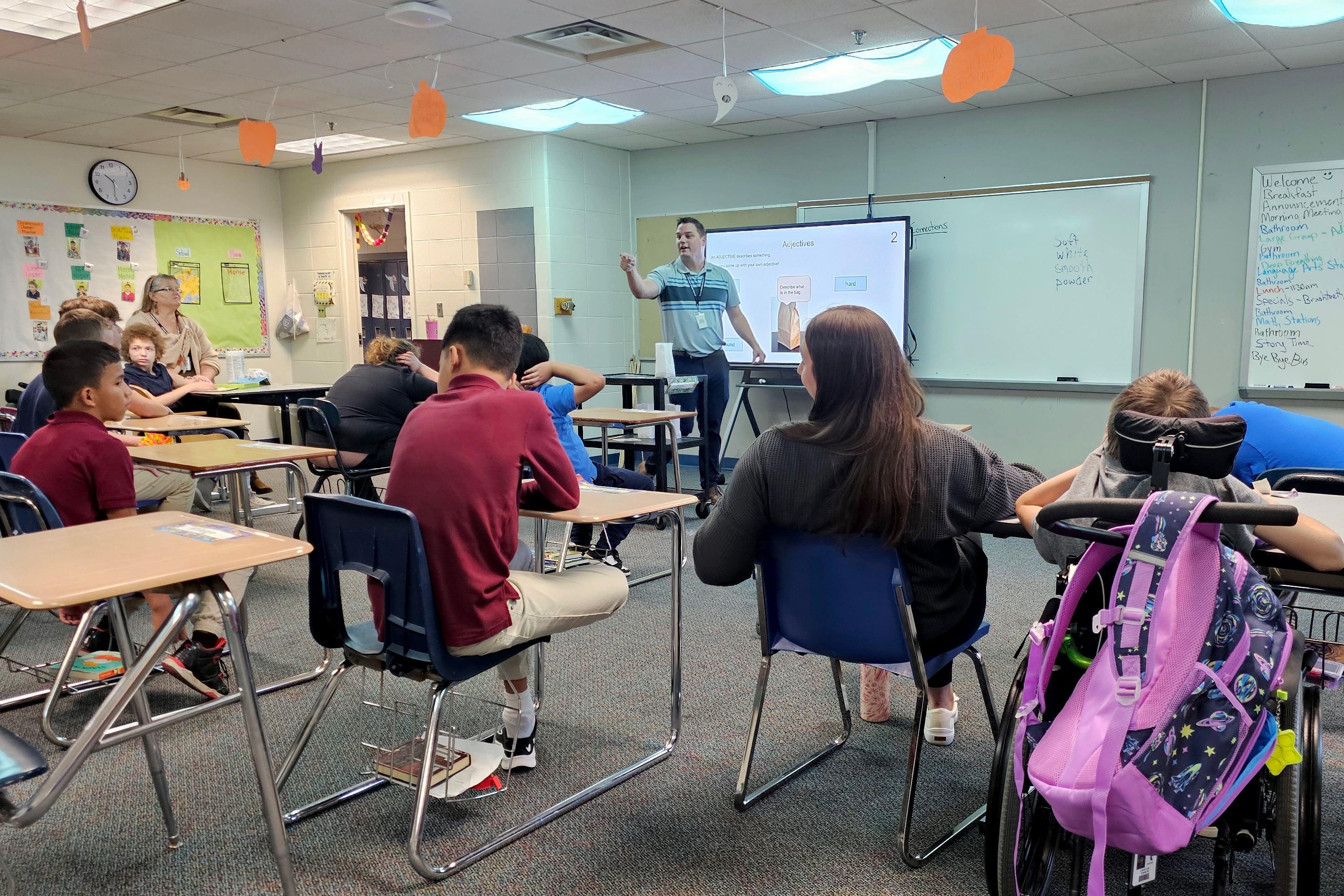 An adult man wearing a blue shirt teaches from the front of a classroom to a room full of students sitting at desks.