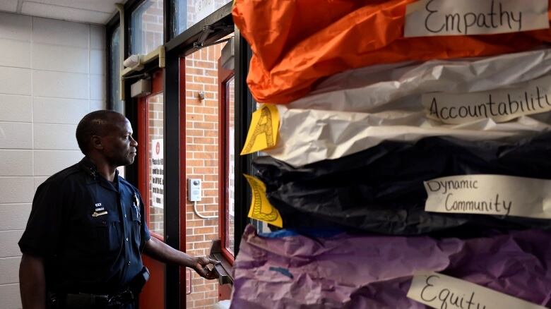 A Black man in a police uniform looks out of a school door that he’s holding slightly ajar. On the right side of the image, there’s colorful paper with labels attached that read: Empathy, Accountability, Dynamic Community, and Equity.