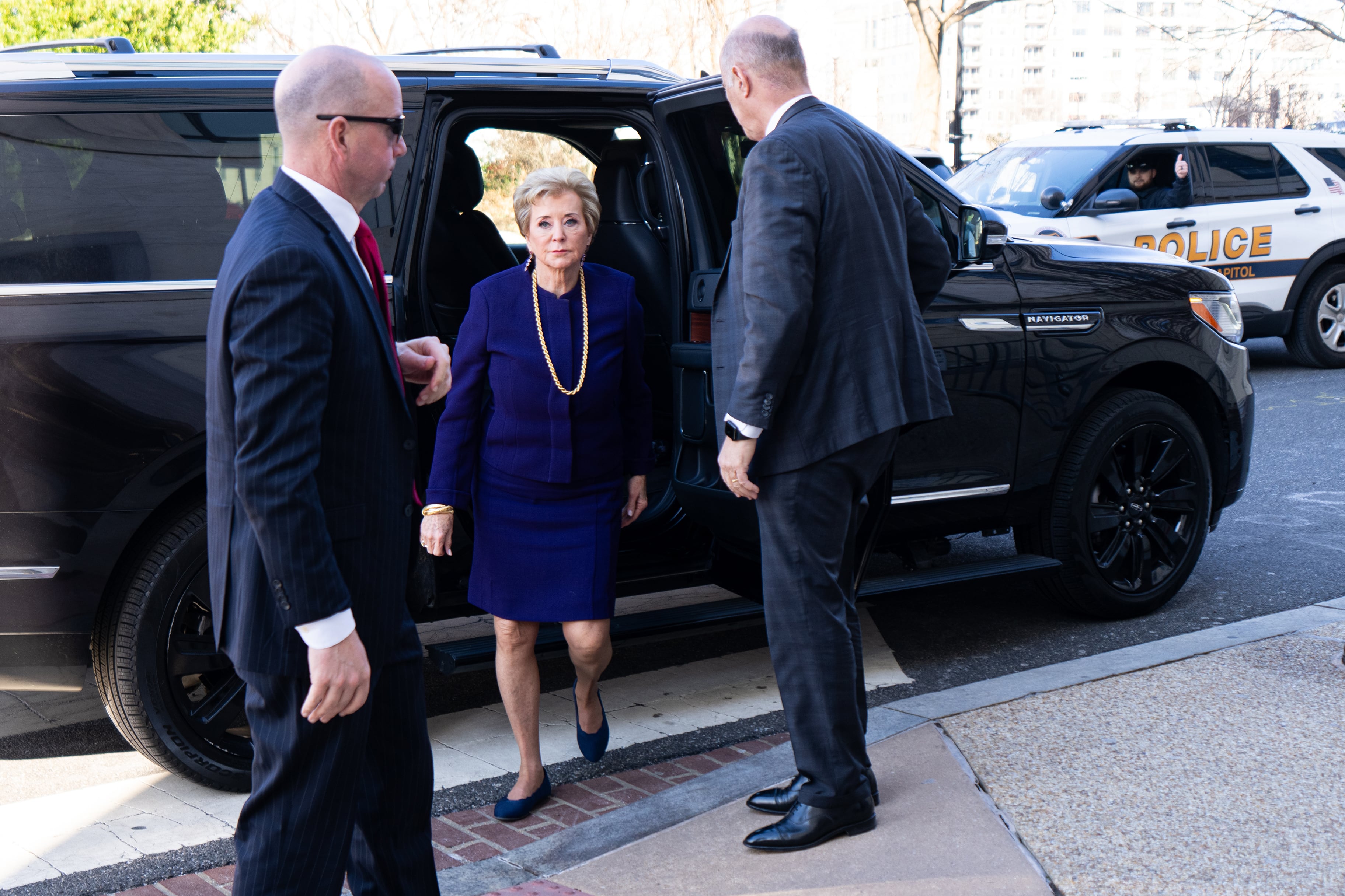 A woman in a purple dress and a long, gold chain gets out of a black SUV with two men in suits on either side of her.