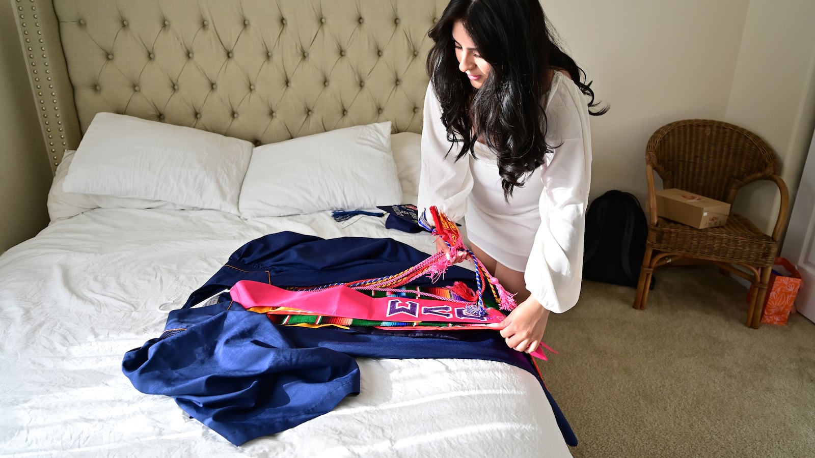 A young woman prepares her blue graduation gown for a ceremony. She has long dark hair, and places sashes over the gown on her bed.