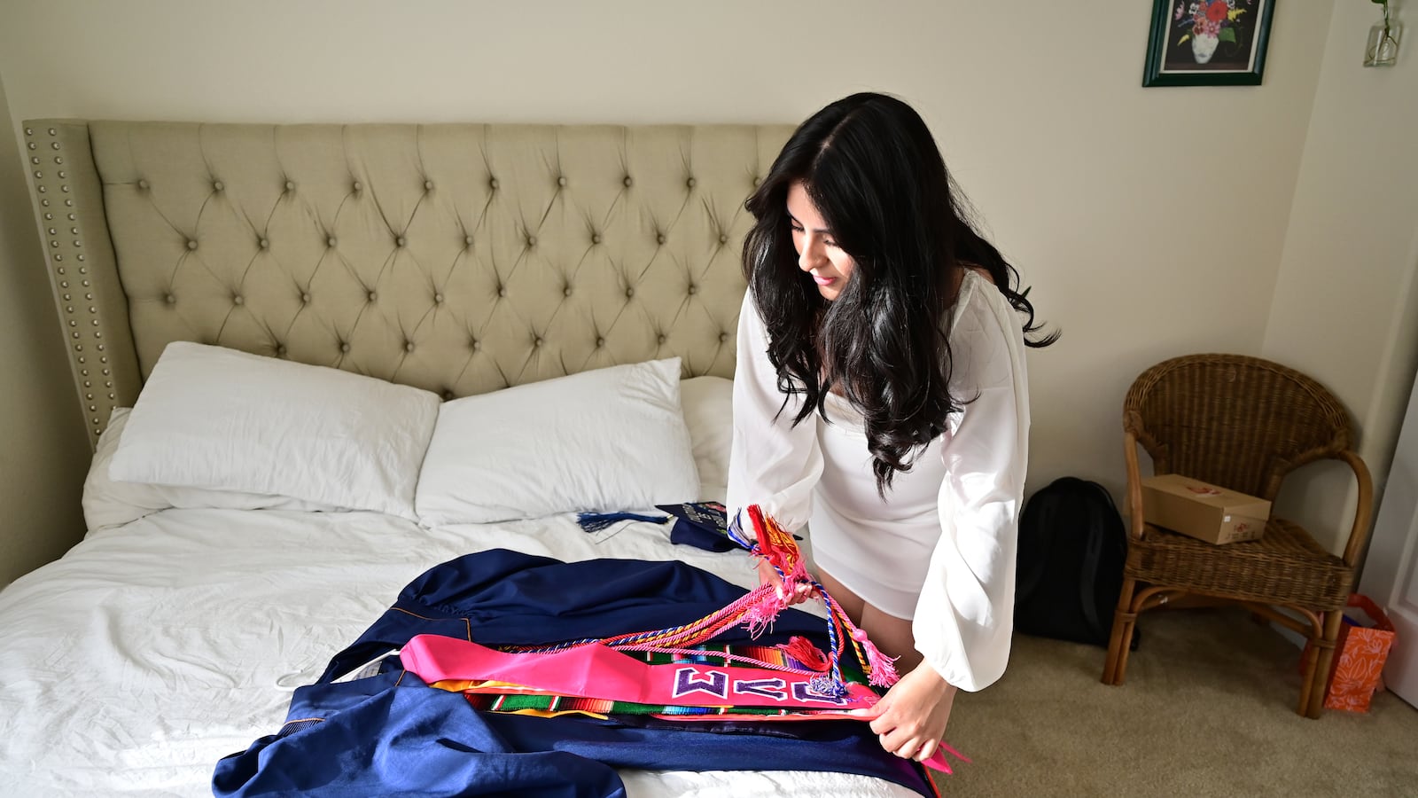 A young woman prepares her blue graduation gown for a ceremony. She has long dark hair, and places sashes over the gown on her bed.