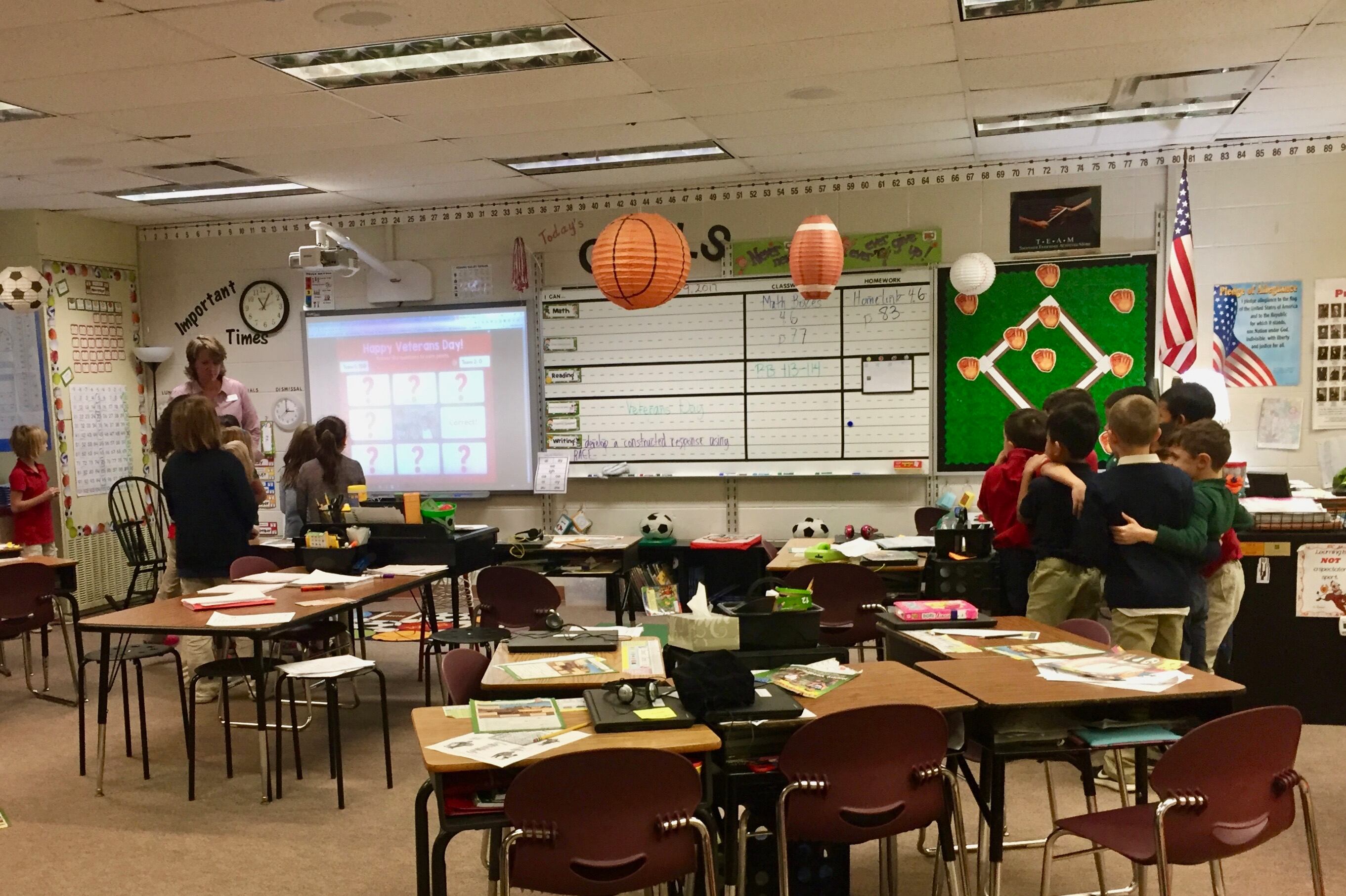 A group of elementary students stand huddled on one side of a classroom amid desks. On the other side of the classroom, a teacher speaks to another group of students looking at a projector.