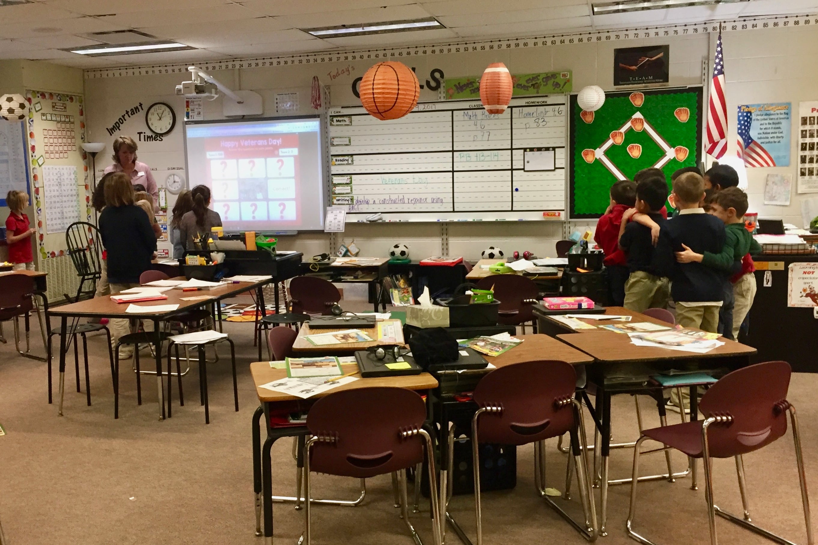 A group of elementary students stand huddled on one side of a classroom amid desks. On the other side of the classroom, a teacher speaks to another group of students looking at a projector.