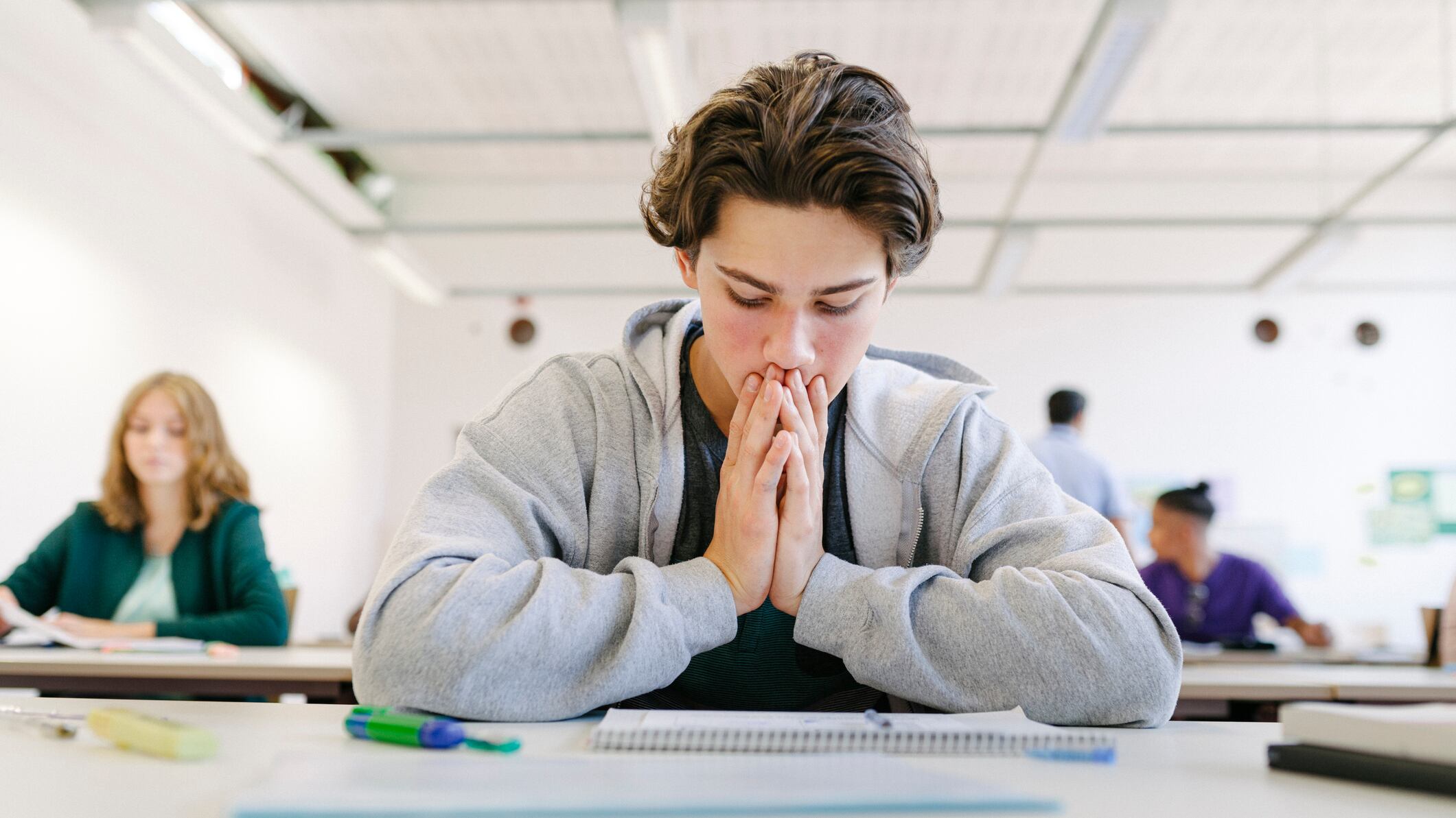 A student in a gray hoodie looks down at a paper exam. His hands are put together and resting against his chin and mouth.