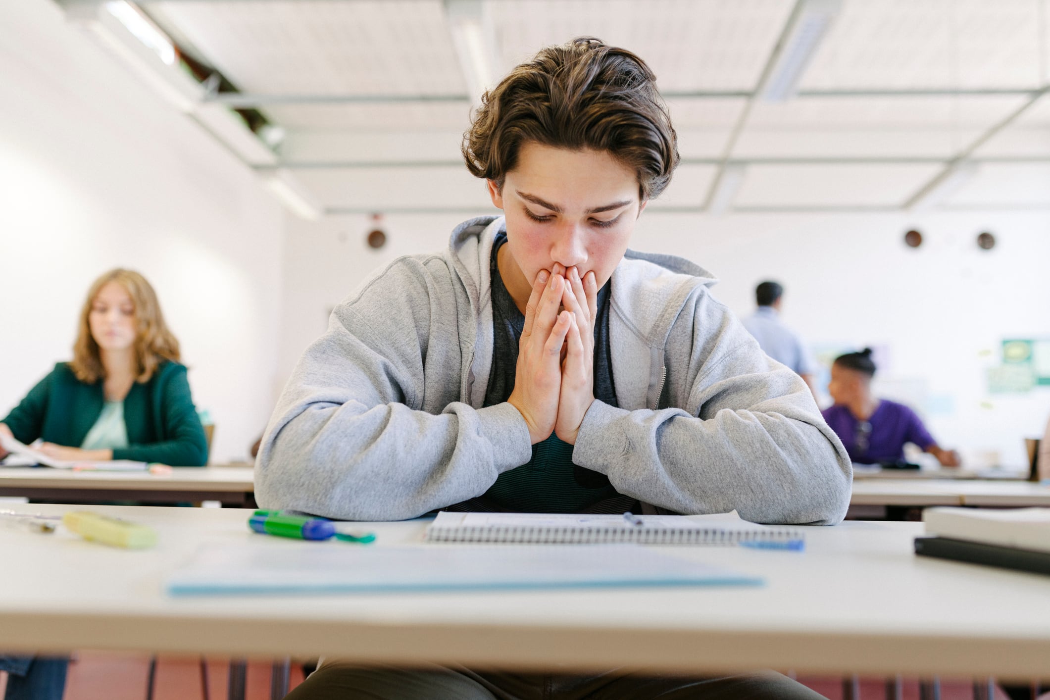 A student in a gray hoodie looks down at a paper exam. His hands are put together and resting against his chin and mouth.