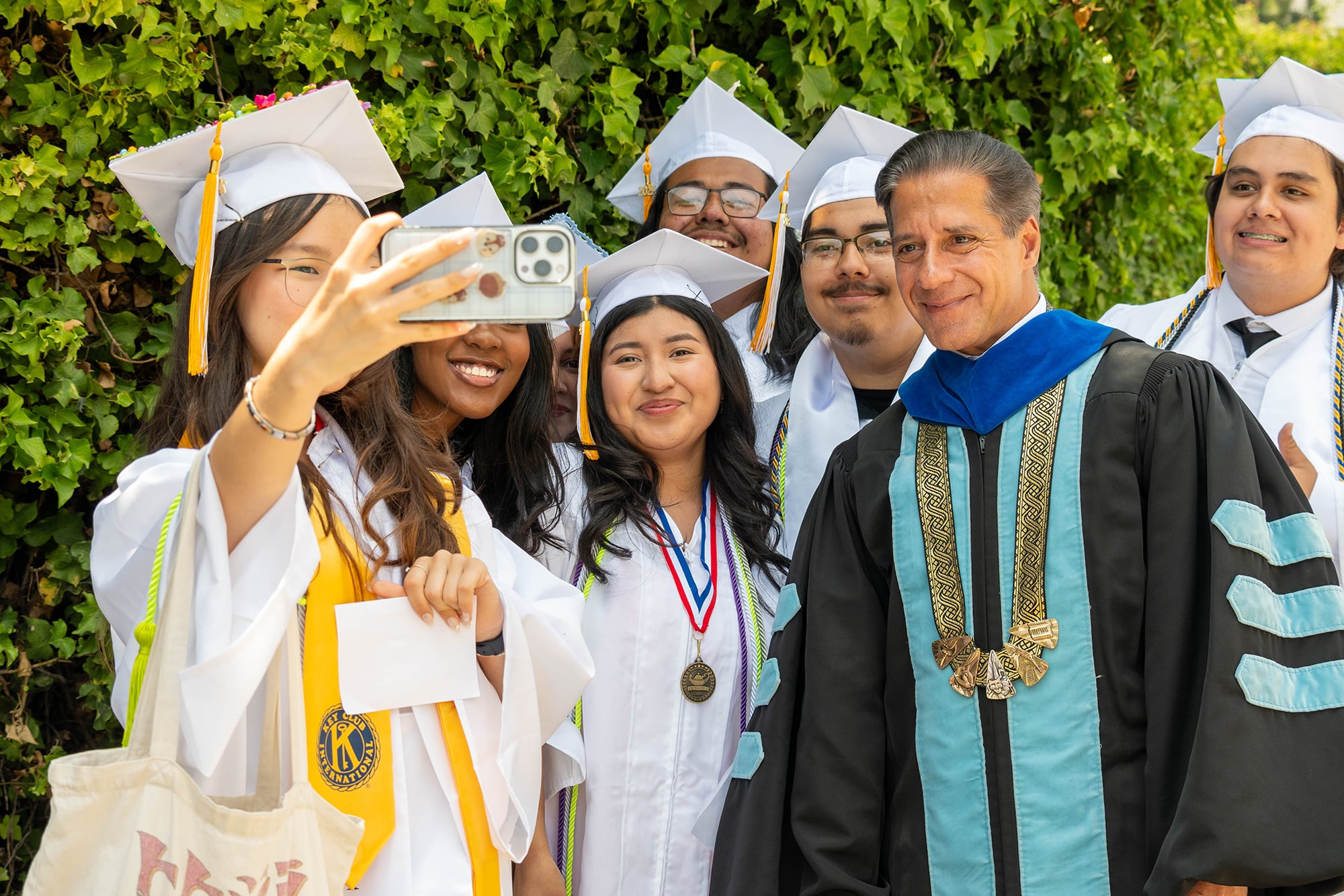 Six high school graduates pose for a photograph with an adult outside on a sunny day.