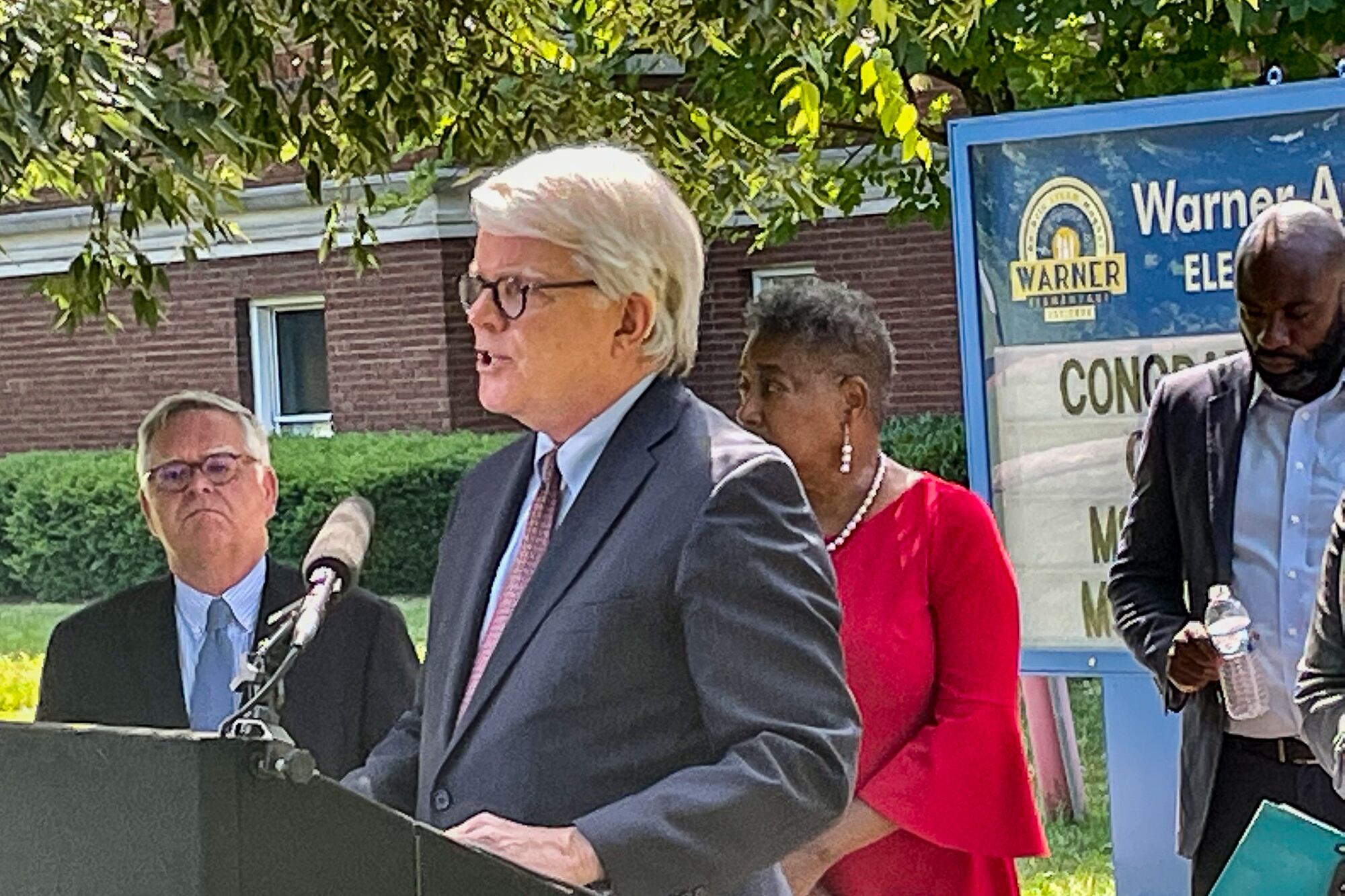 A man wearing a suit speaks at a podium in front of a school and surrounded by several other people wearing suits and dresses.