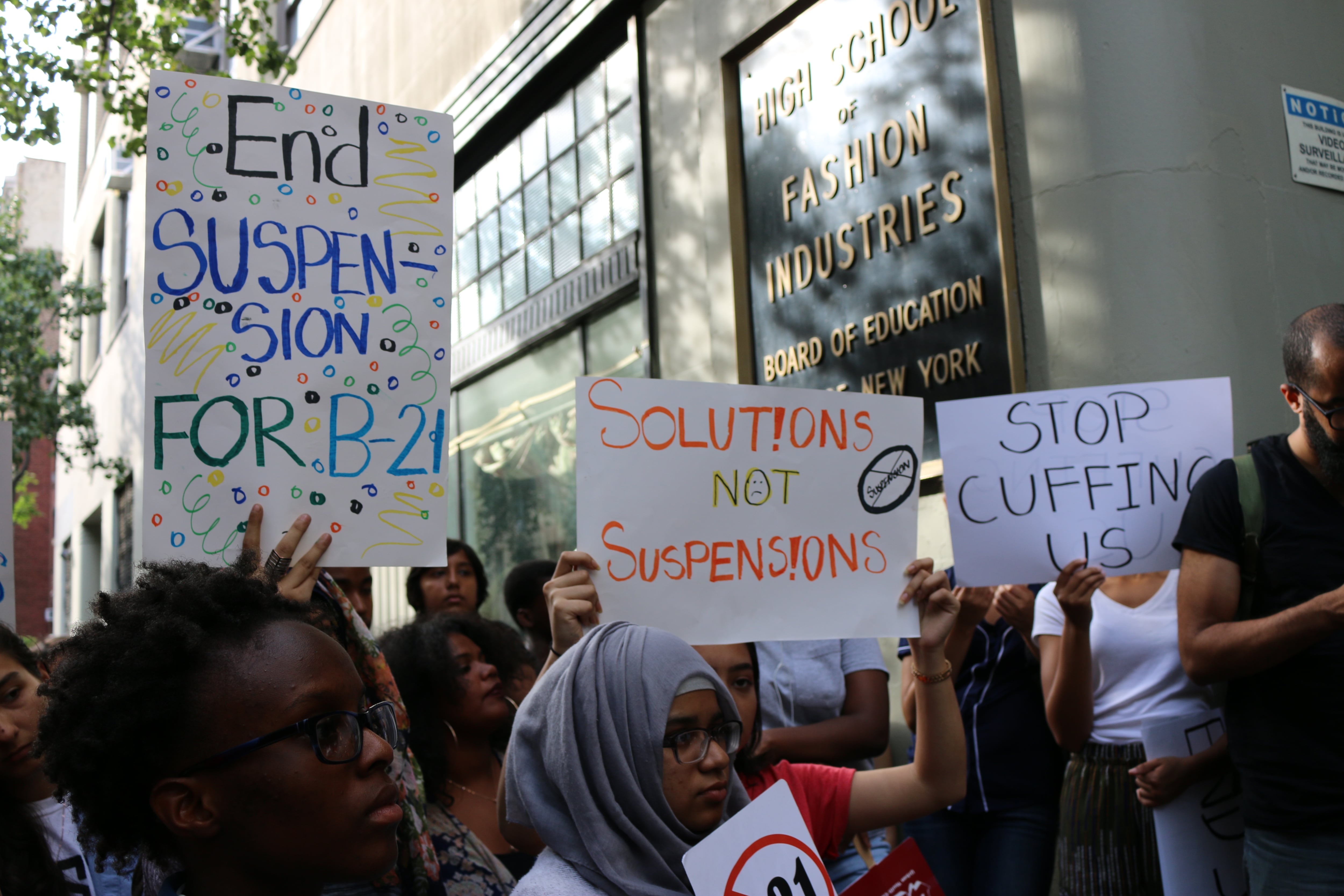 A crowd of people protest outside of the Board of Education building while some people hold up signs that read "solutions not suspensions," and "stop cuffing us."