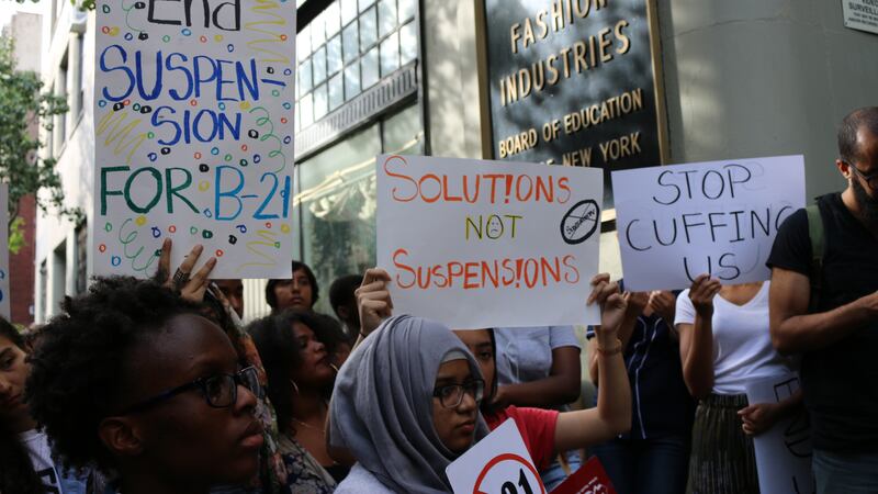 A crowd of people protest outside of the Board of Education building while some people hold up signs that read "solutions not suspensions," and "stop cuffing us."