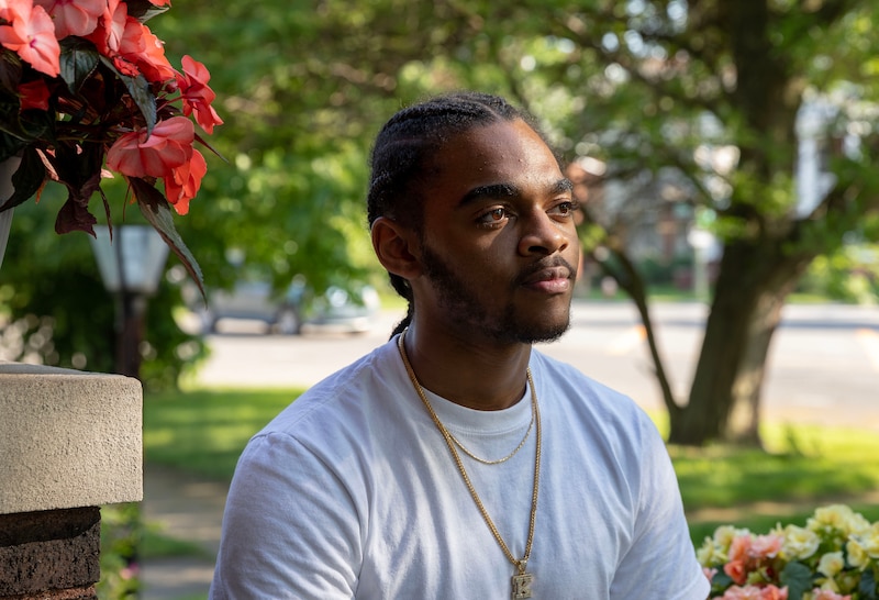 A high school senior with dark hair and wearing a white t-shirt with a gold necklace with a diamond letter "K" poses for a portrait in front of a tree and stree.