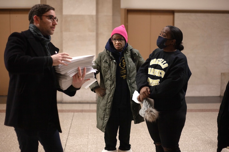 Three people wearing winter coats stand while talking and holding papers with a tan stone wall in the background.