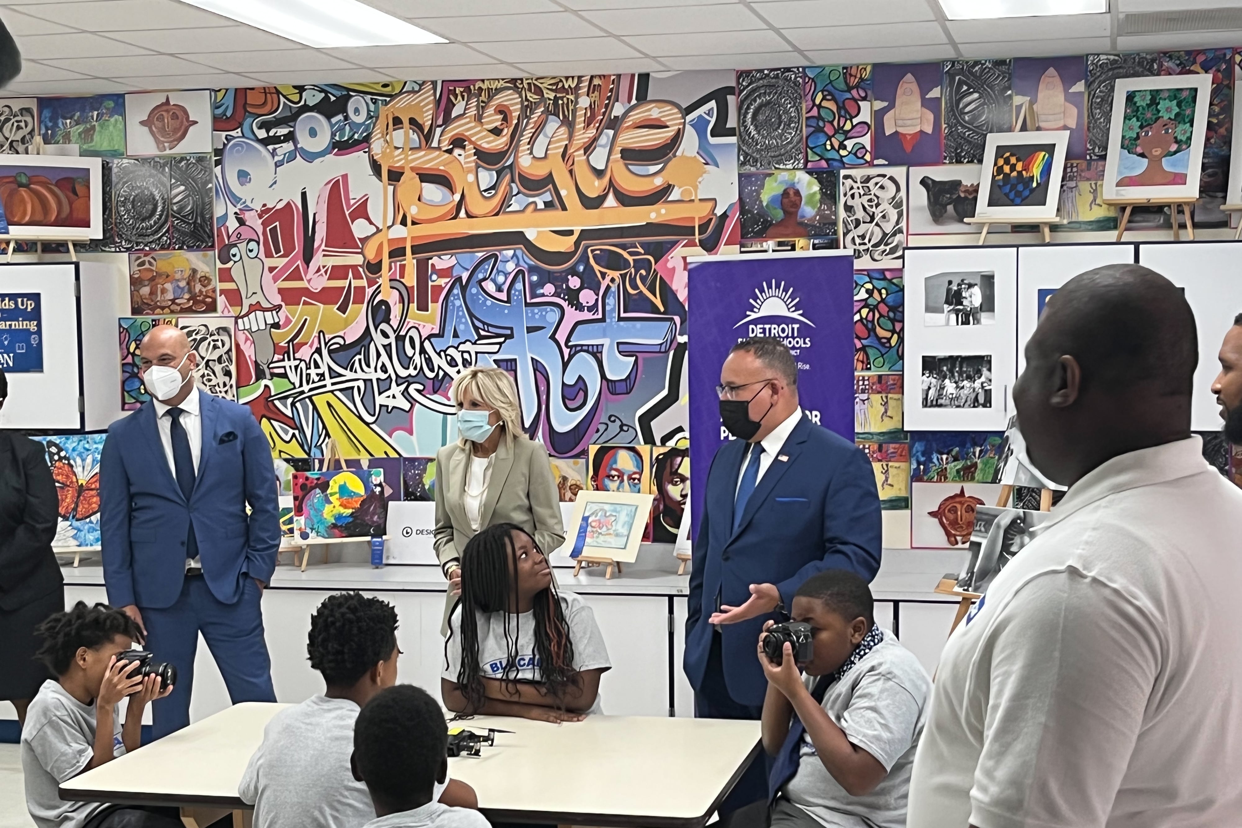 An older man, U.S. Education Secretary Miguel Cardona, stands before a classroom of students to ask them how they are liking their photography lesson. He has his hand gesturing in front of him. To his far left, another older man, Superintendent Nikolai Vitti, stands with his left hand in his pocket. Right beside Cardona, an older woman, First Lady Jill Biden, stands. Two of the students in the classroom are holding cameras.