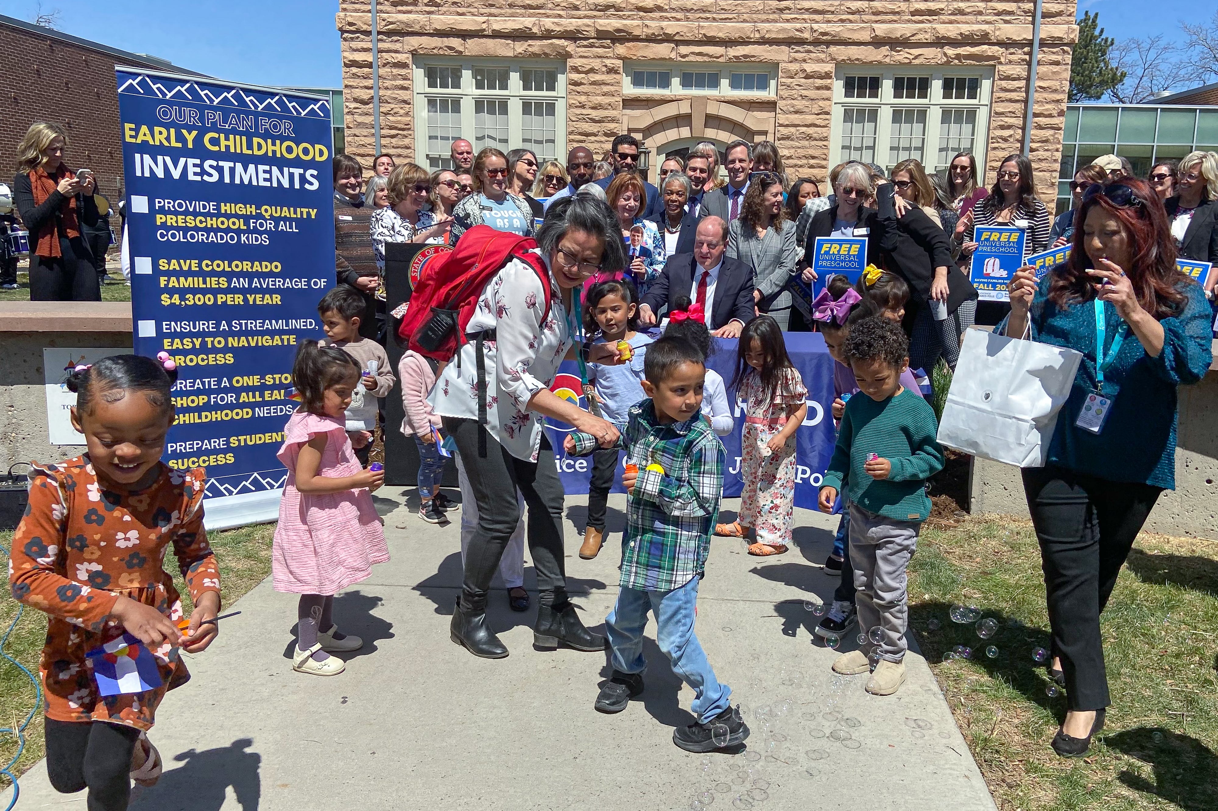 Small children run around chasing bubbles and waving flags in front of Colorado Gov. Jared Polis, who is seated at a table and signing a bill. A large crowd of cheering adults stands behind him.