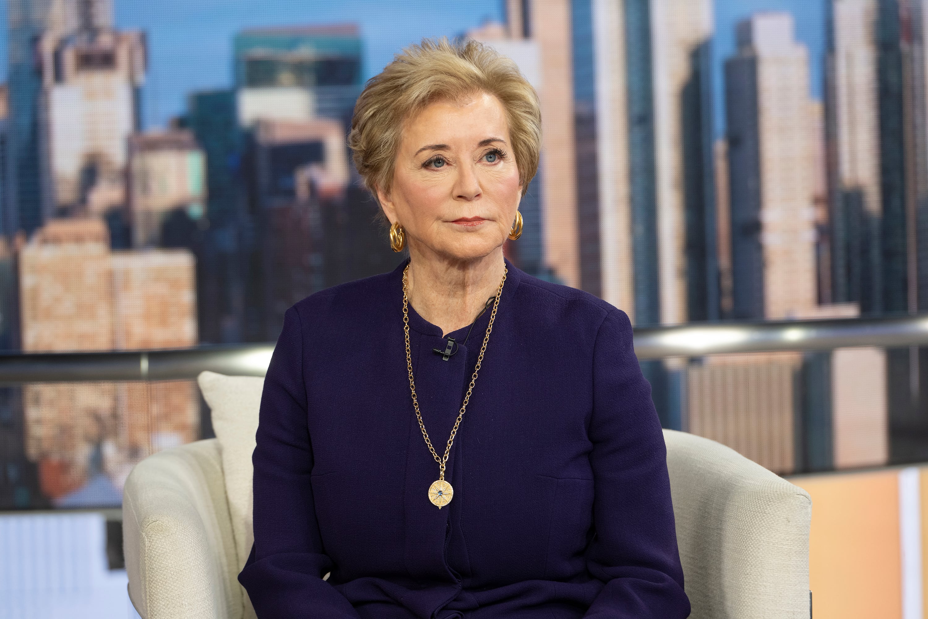 A white woman with short blonde hair and wearing a dark blouse sits in a chair with a cityscape behind her.
