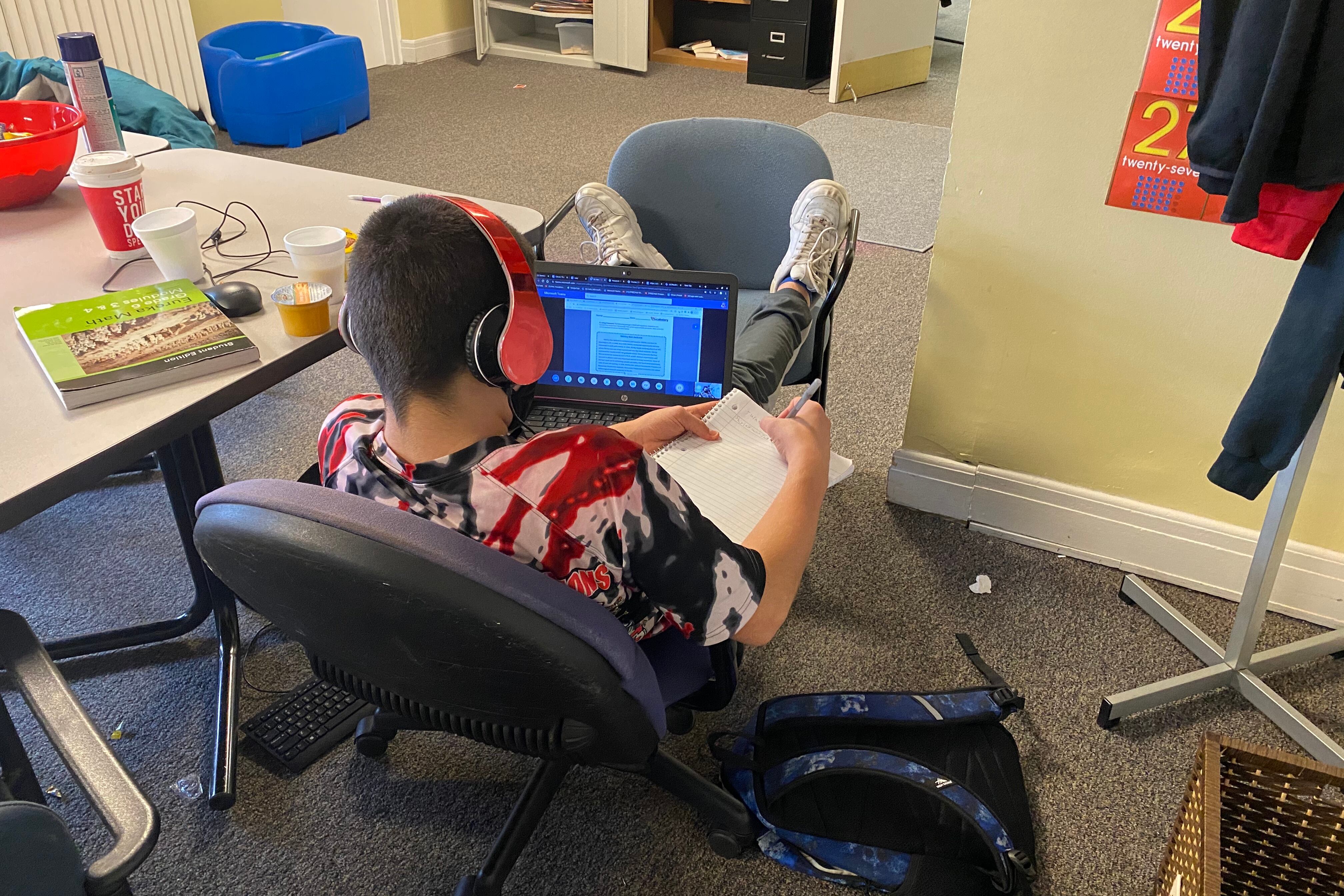 A student with headphones on sits in a chair writing in a notebook with a computer on his lap in a room.