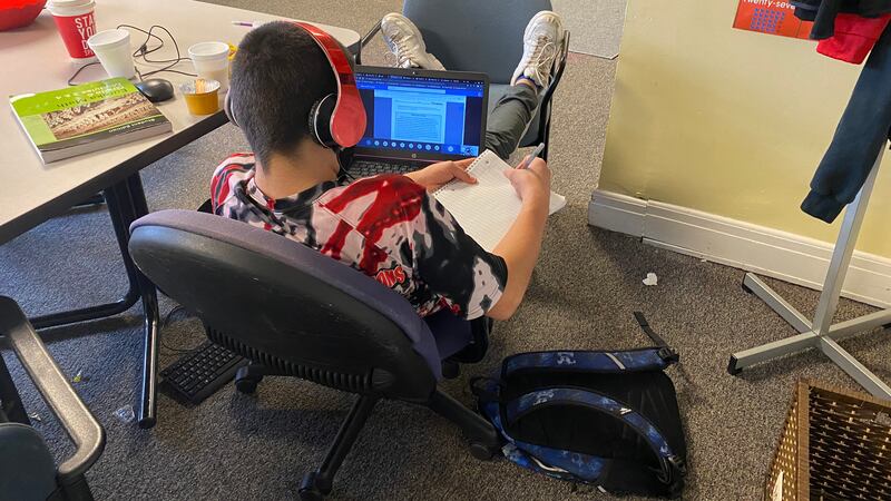 A student with headphones on sits in a chair writing in a notebook with a computer on his lap in a room.
