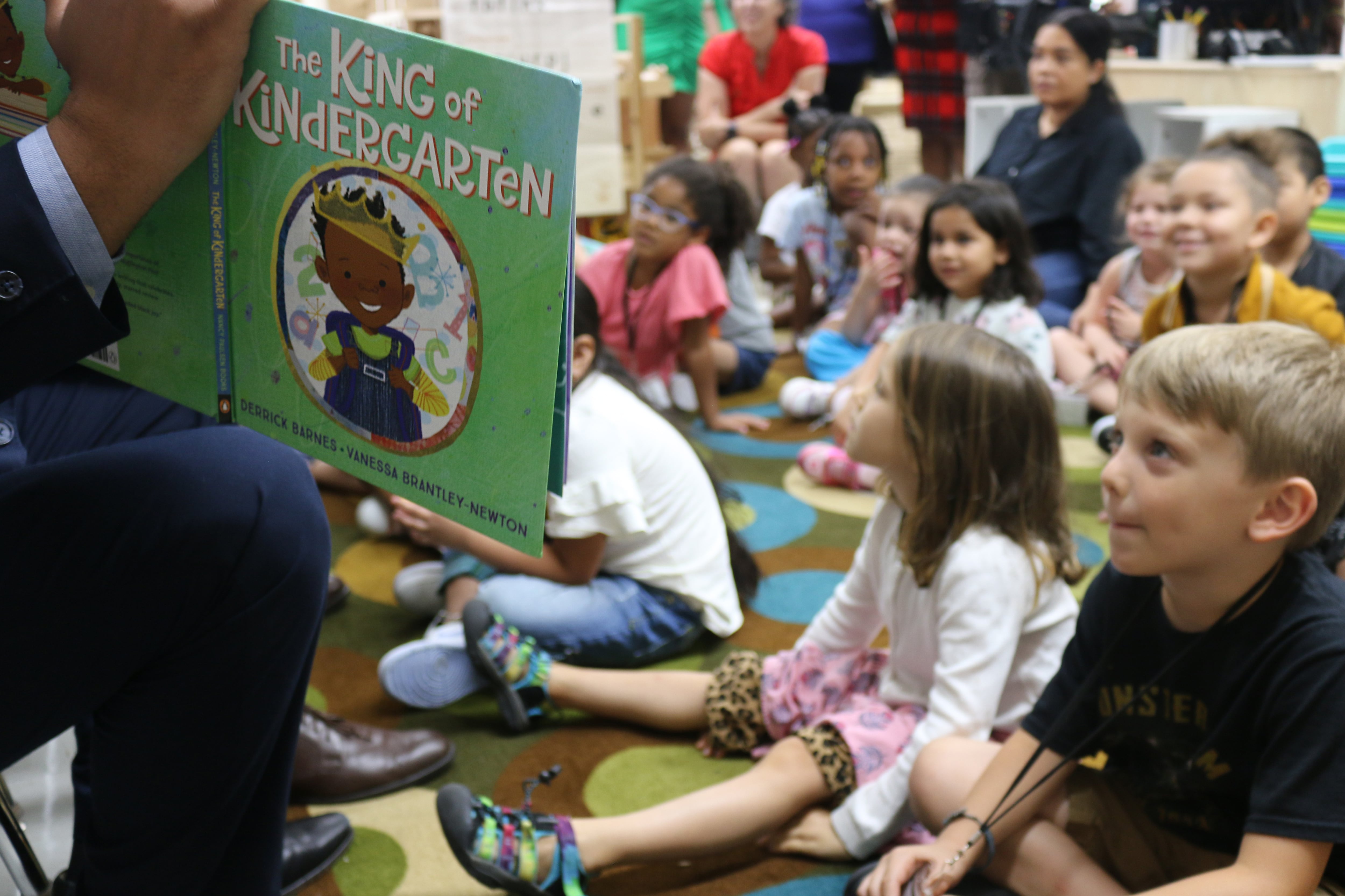 A hand holding a book in the foreground of a large group of students sitting on a rug in a classroom.