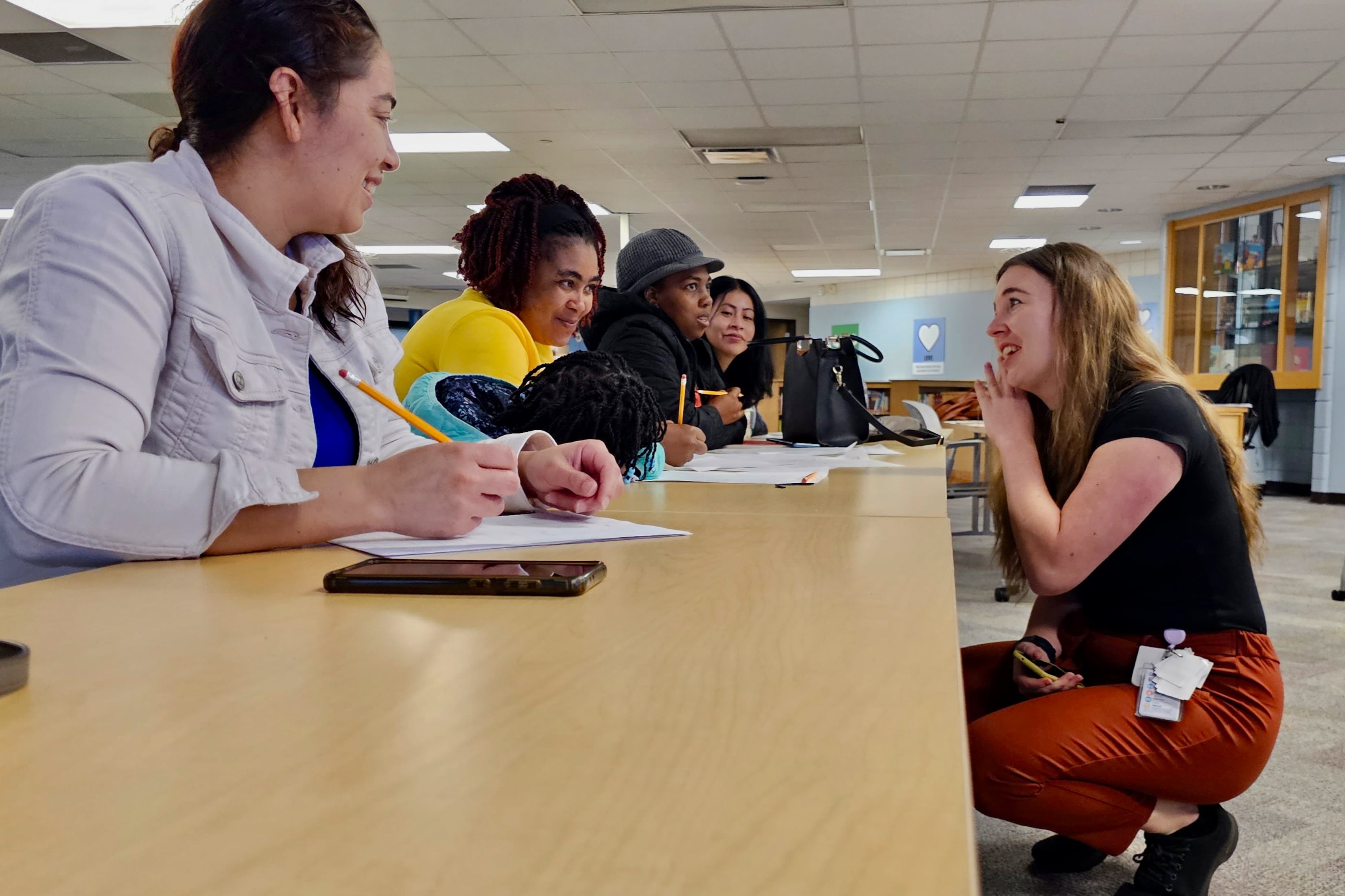 A teacher in a black top and red pants kneels in front of a desk where four adult students are writing on paper with pencils. Between two of the adults, a child in a blue jacket has her head down on the desk.