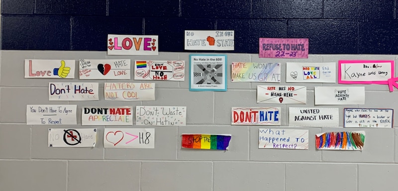 A group of handmade bumper stickers with anti-hate messages are displayed on a black and grey wall.