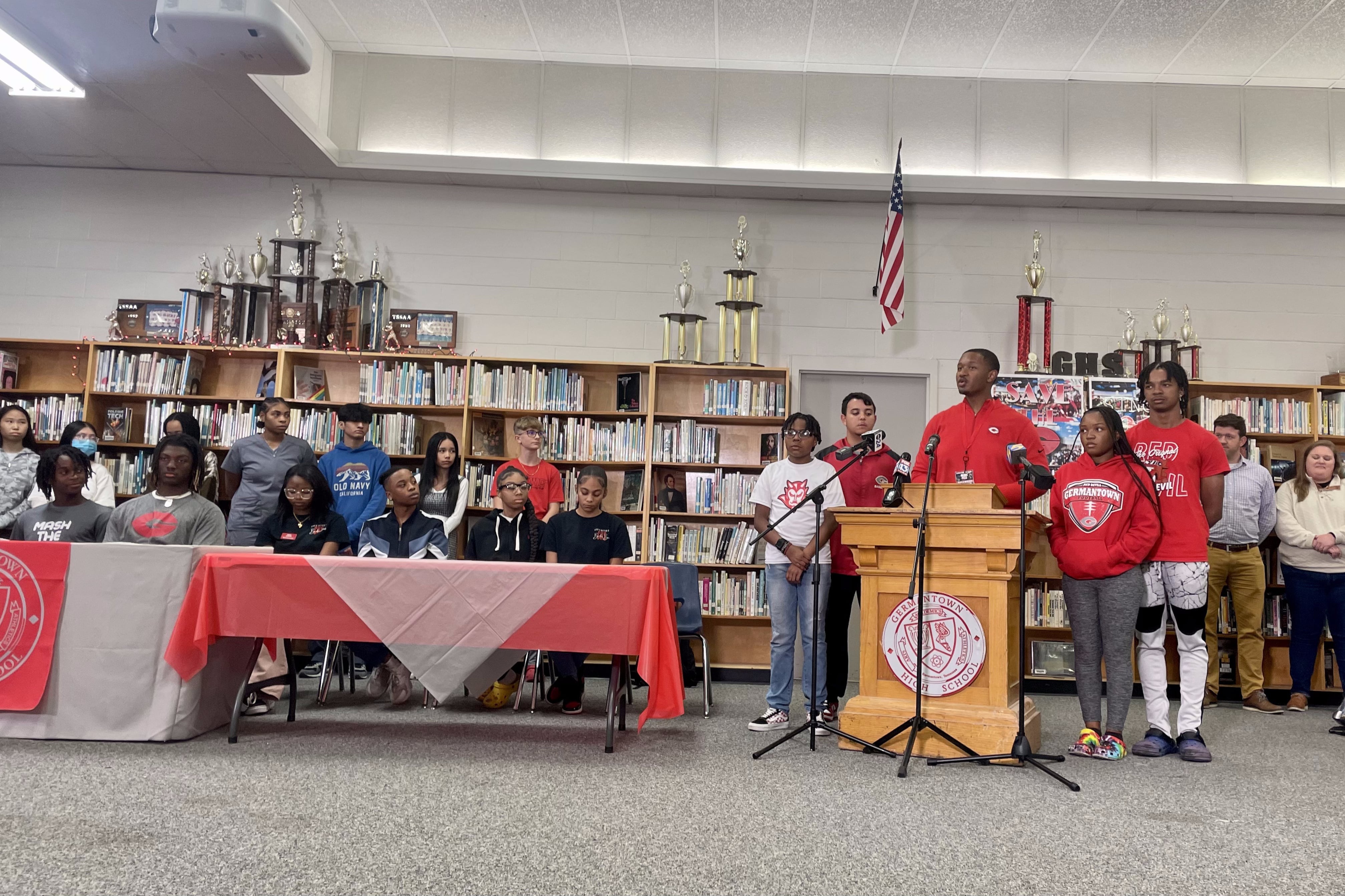 A student speaks at a wooden “Germantown High School” podium in a school library as about 20 other students and teachers look on
