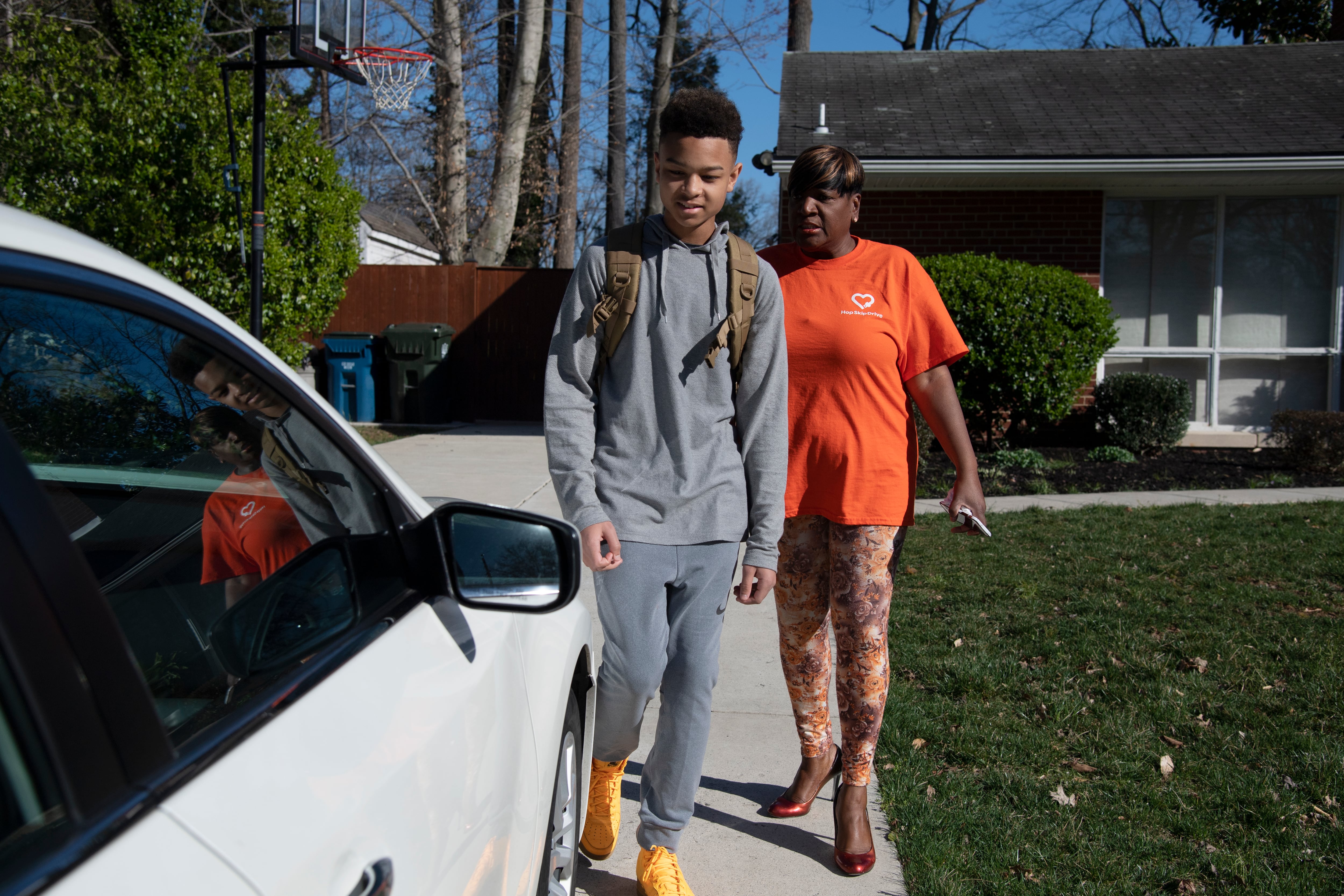 A teen in gray and a woman in an orange top walk on a driveway.