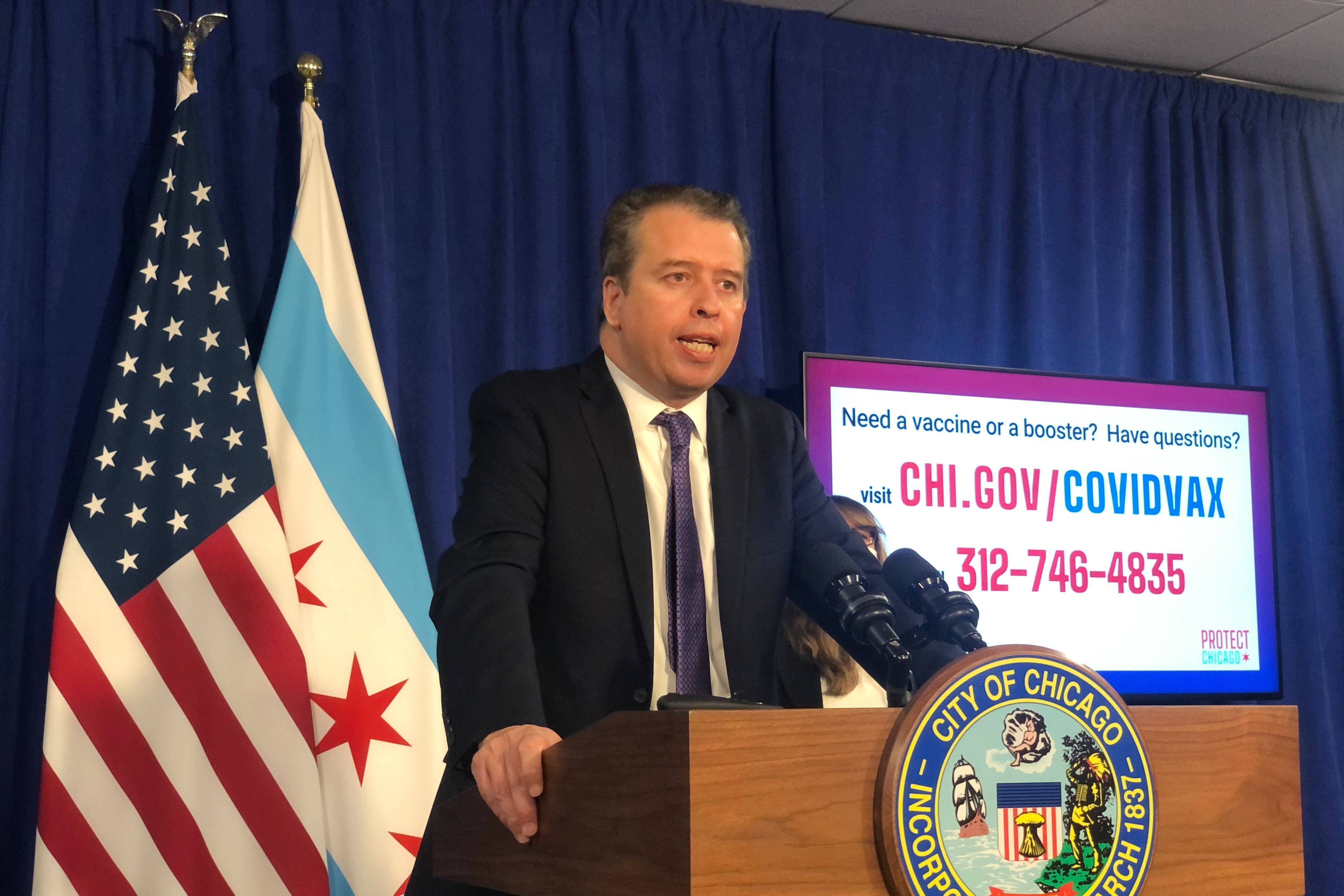 Chicago Public Schools CEO Pedro Martinez, wearing a black suit and purple tie, speaks at a wooden City of Chicago podium during a press conference. There are Chicago and United States flags hanging behind him against a blue backdrop, along with a screen providing COVID information.
