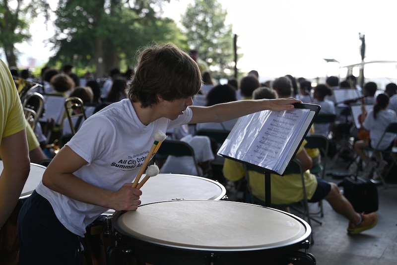 A photograph of a male high school student flipping the page of sheet music standing in front of a large drum with a view of students playing instruments in the background.