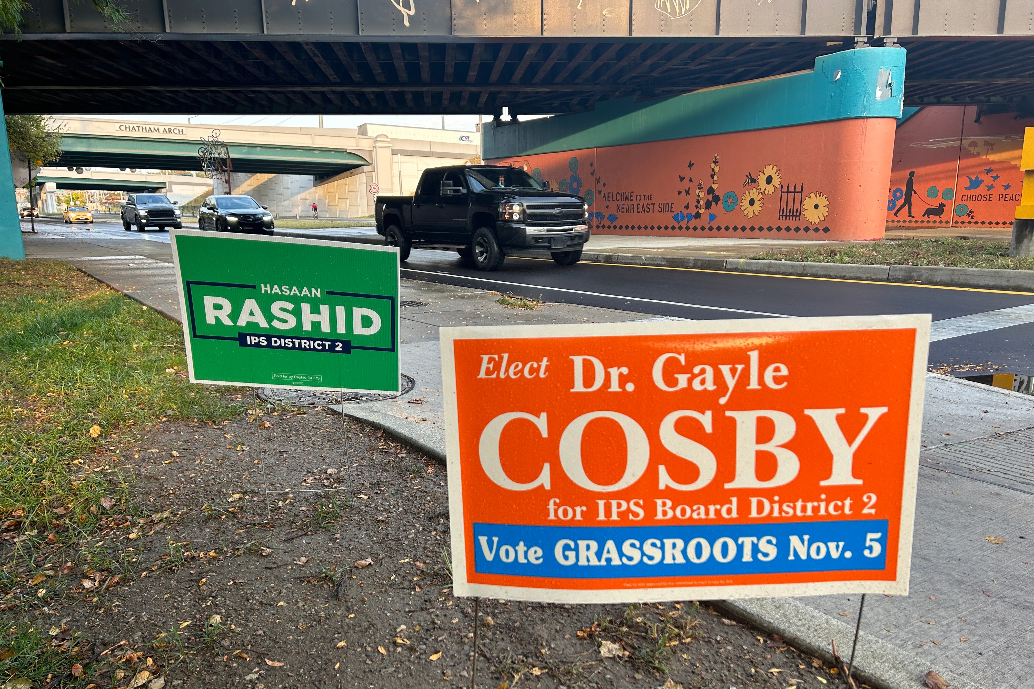 Two campaign signs, one green and one orange, sit by a street with cars passing by. The green sign reads "Hasaan Rashid District 2." The orange sign reads "Elect Dr. Gayle Cosby for IPS Board District 2. Vote grassroots Nov. 5."