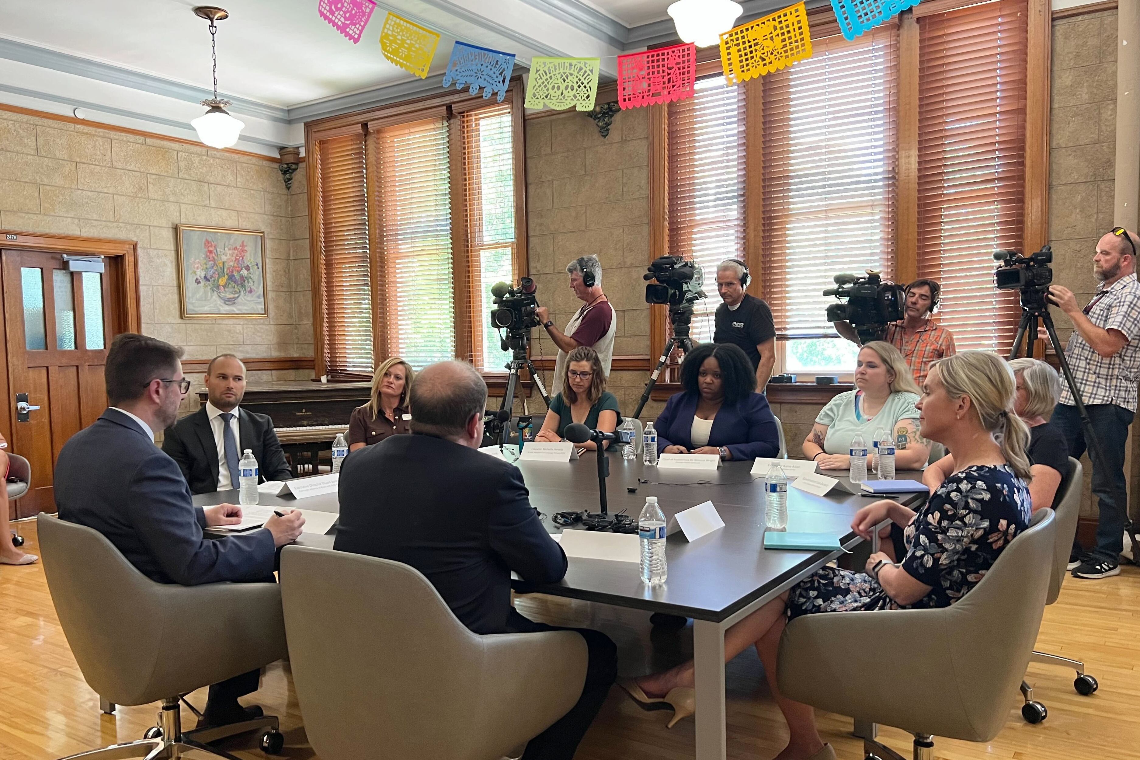 A group of adults sit around a table while a row of camera operators in a large room.