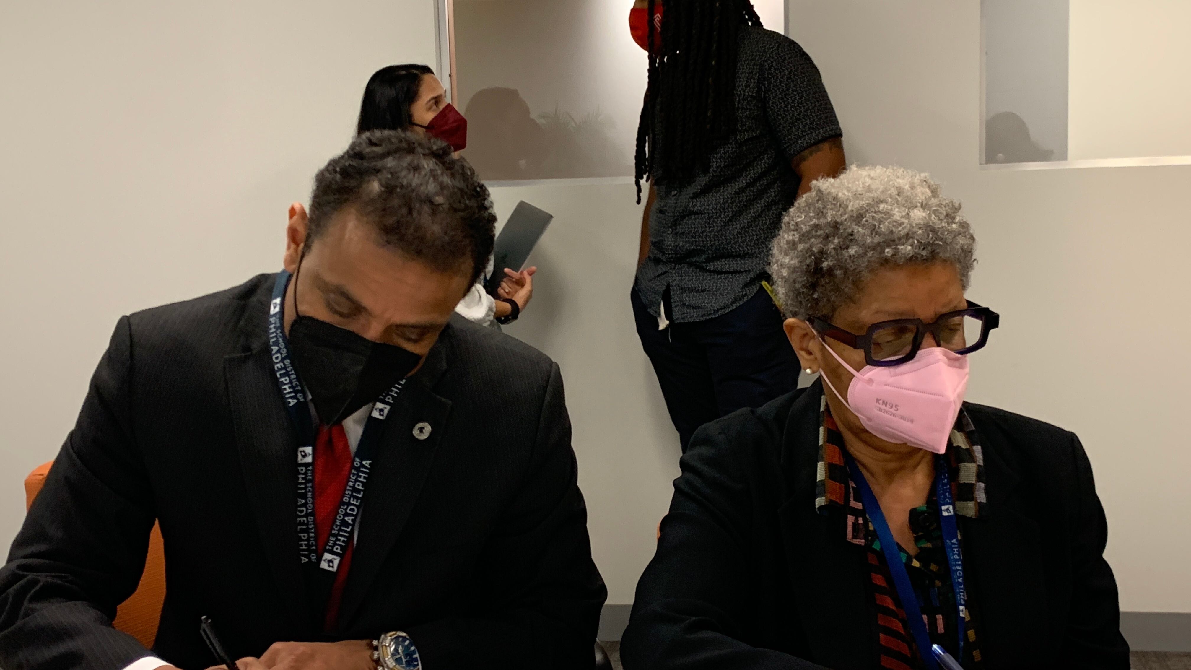 A man and a woman wearing dark tops sit next to each other while wearing masks and sign documents at a table.