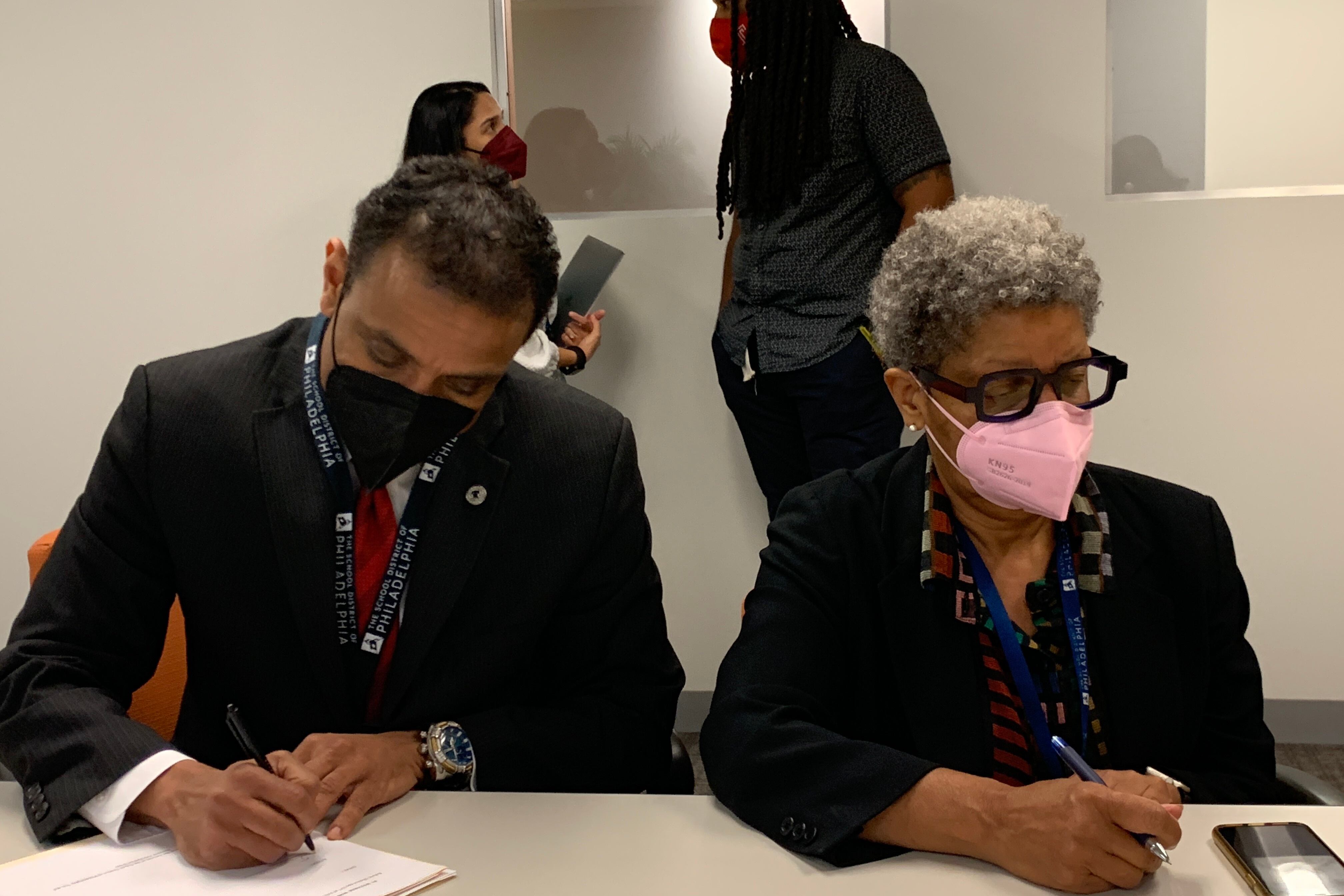 A man and a woman wearing dark tops sit next to each other while wearing masks and sign documents at a table.