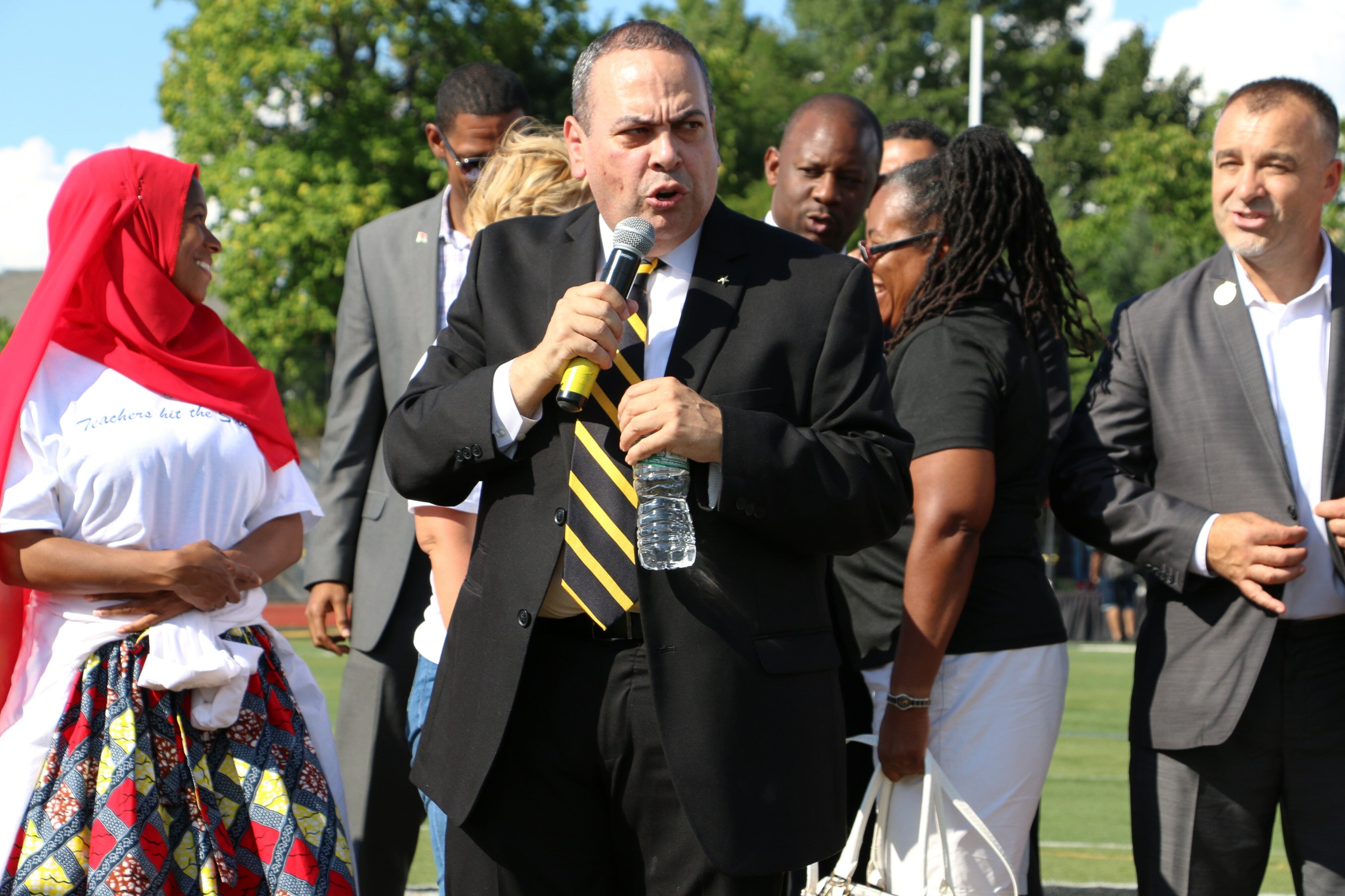 A group of adults in business clothes stand together while one man in a suit speaks from a microphone.