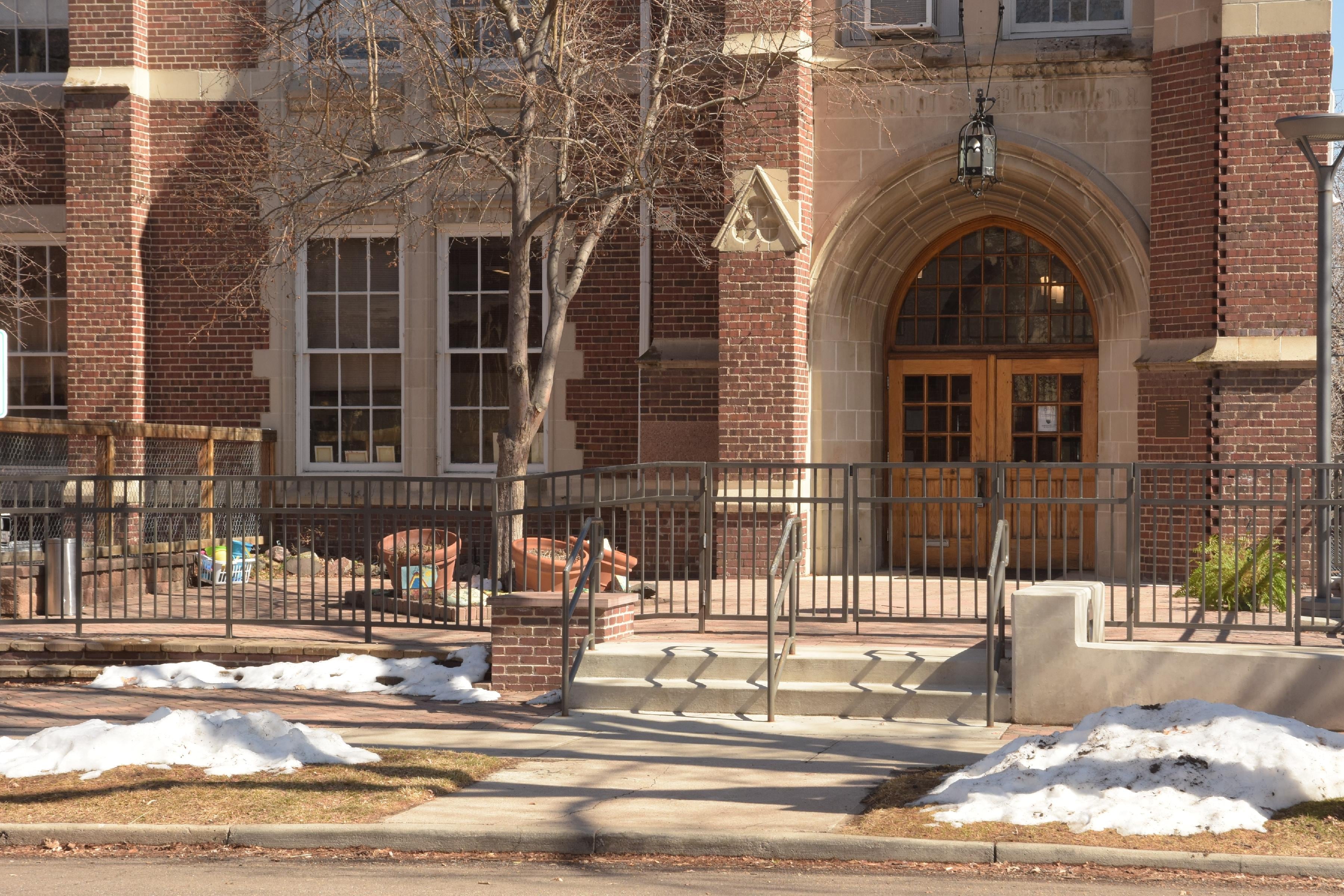 The building of REACH Charter School in Denver. Its wooden doors sit behind a metal fence, and its facade has large windows and red brick walls.