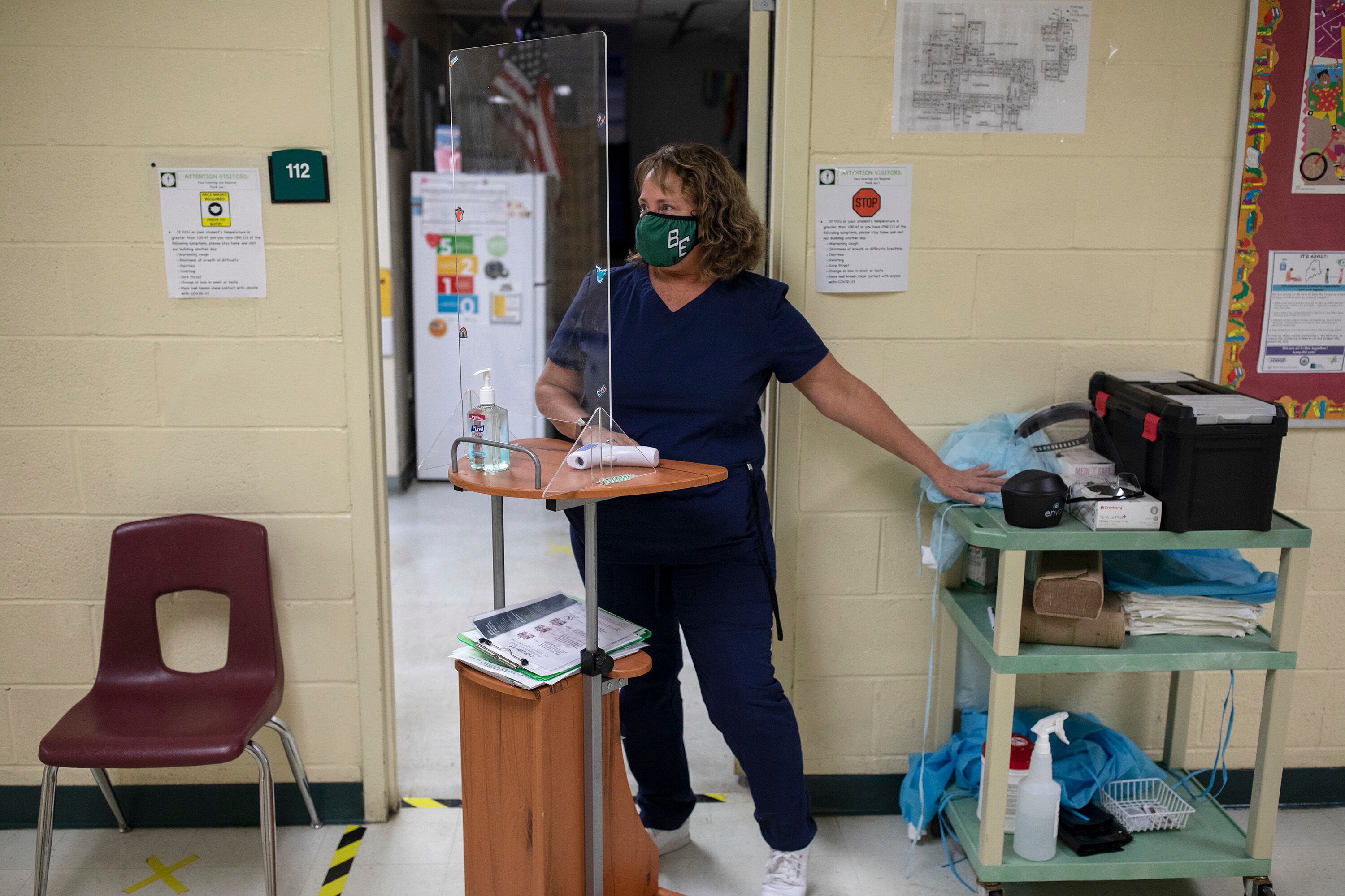A masked school nurse in a uniform stands behind a clear plastic partition and a cart holding an instant-read thermometer and hand sanitizer, and next to a larger cart with cleaning supplies near the open door of an office.