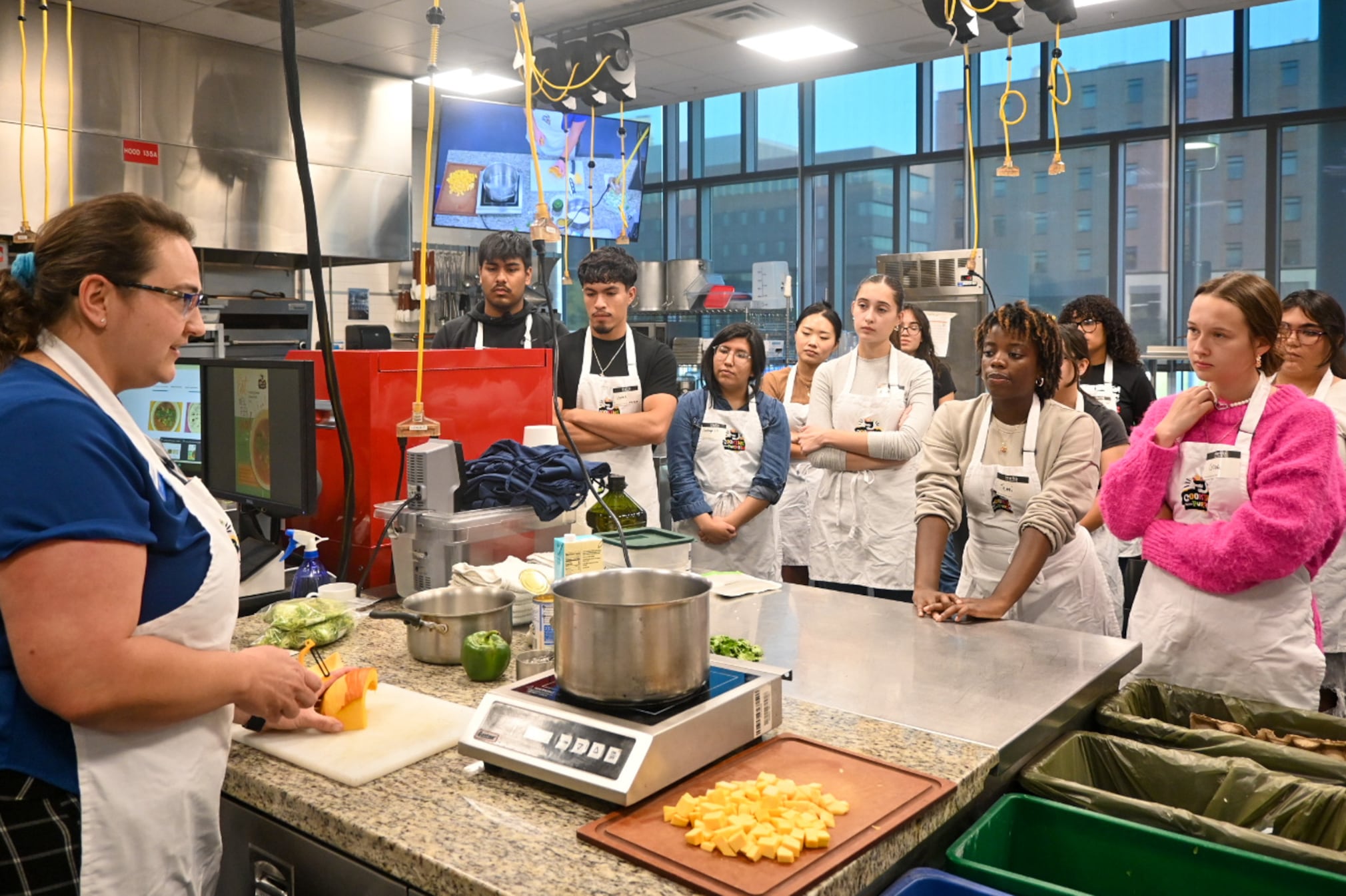 A woman in blue and an apron works with a squash in front of a bunch of students with aprons.