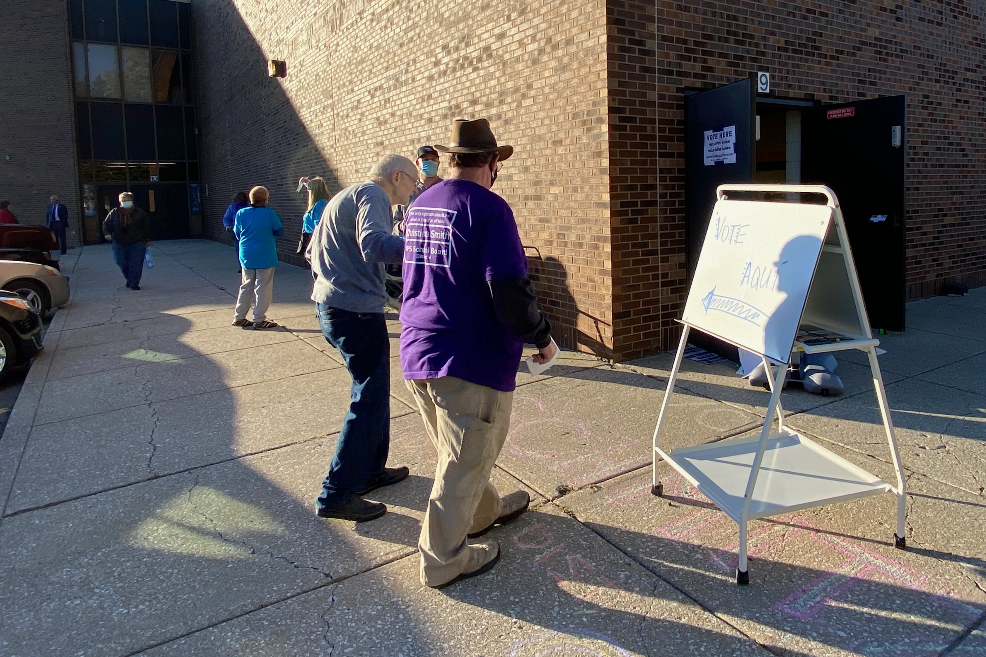A campaign worker helps walk a man past a sign on an easel on a patio outside a polling station.