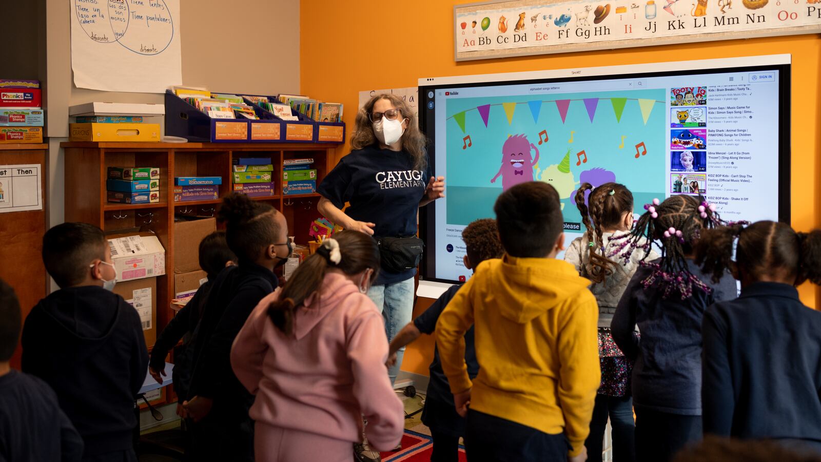 A teacher, wearing a filter mask and navy blue Cayuga Elementary school shirt, leads her young students in an active exercise during class.