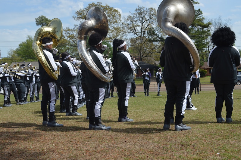 A marching band performs outside with students and trees in the background.