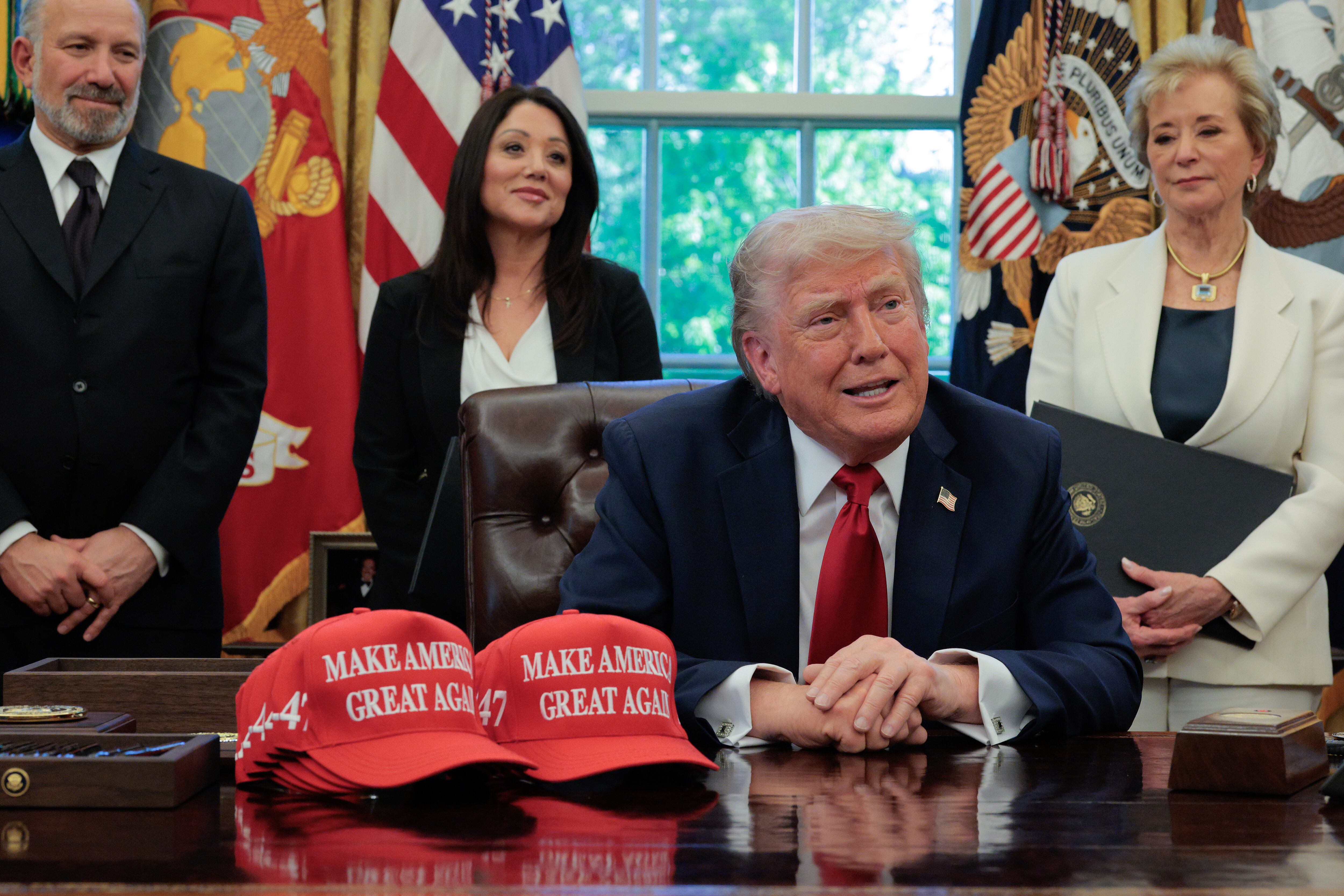 Donald Trump sits at his desk with two MAGA hats surrounded by members of h is cabinet.