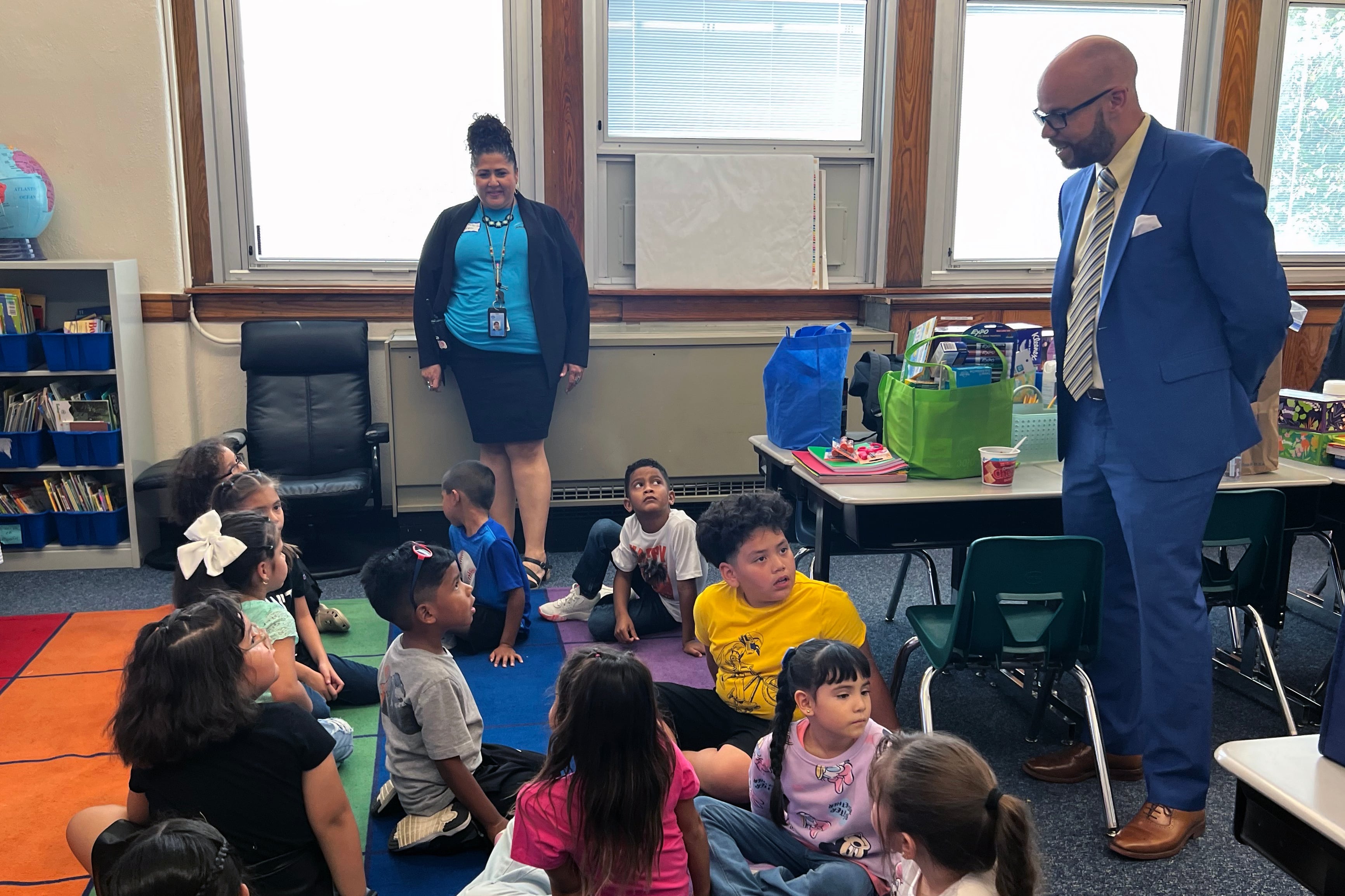 Two people stand next to a group of young students sitting on a colorful rug in a classroom.