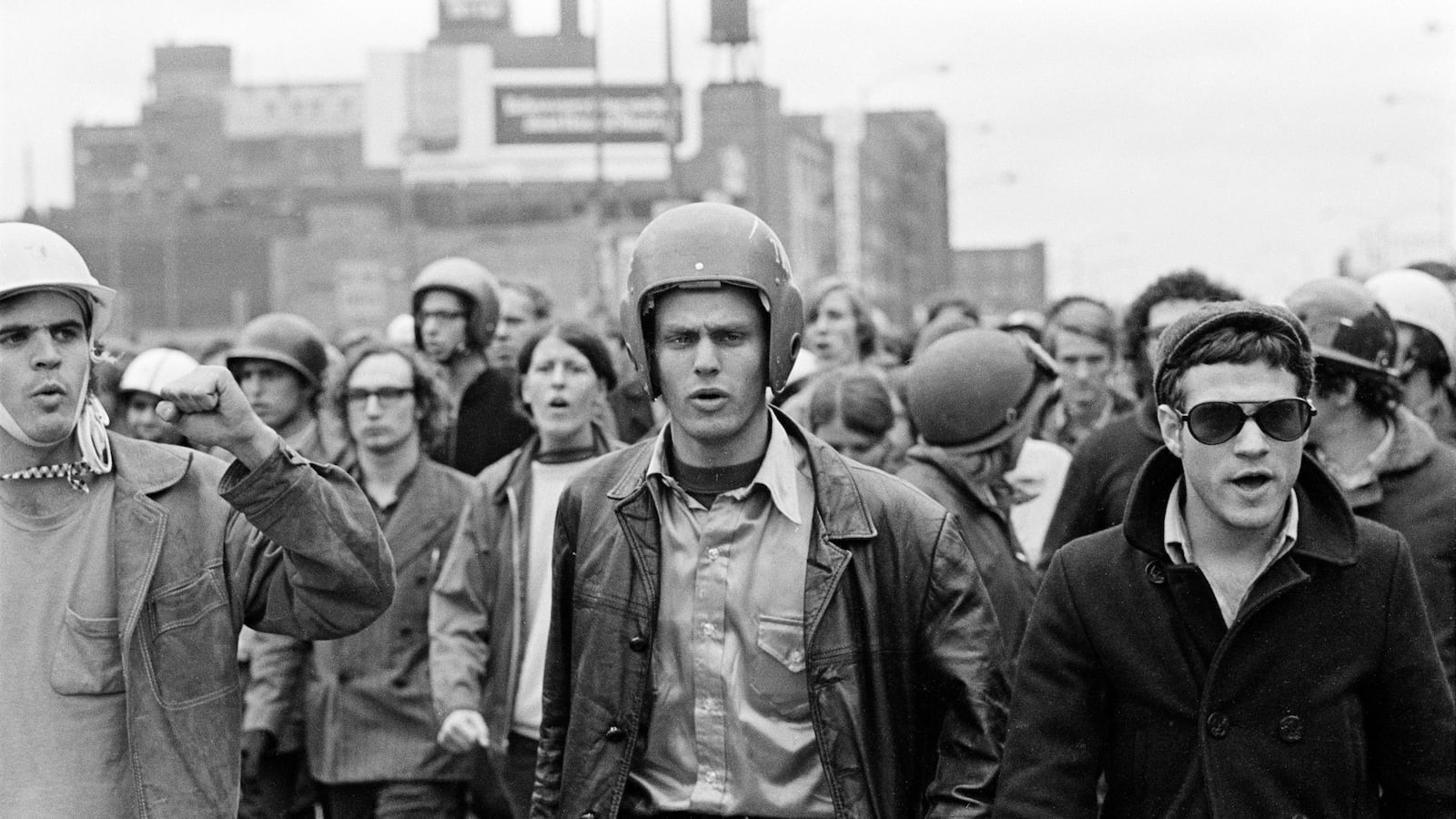 A black and white image of a group of young men in helmets and jackets outside with buildings in the background.