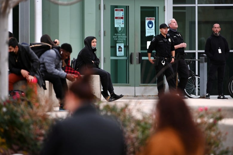Teenagers in hoodies sit on benches outside a school building. Several police officers in uniform stand outside the doors.