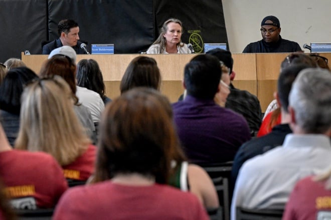 Three school board members sit at a dais during a meeting. The backs of the audience’s heads are visible.