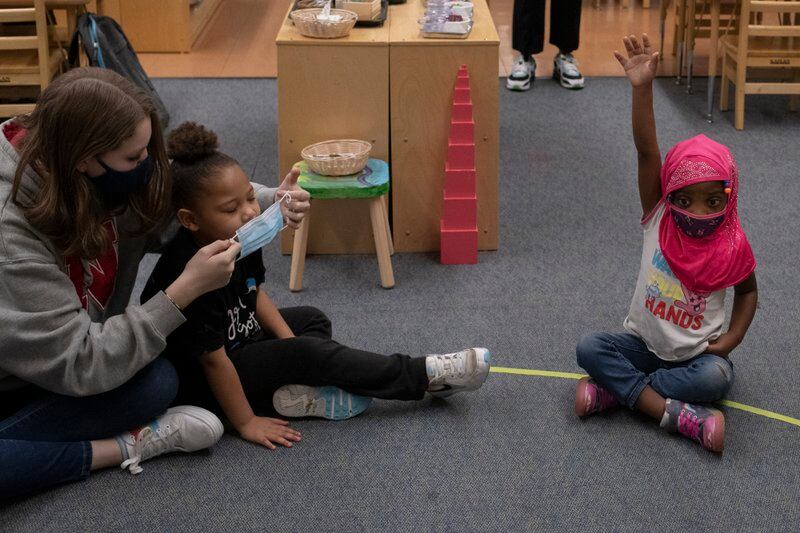 A young girl in a mask and pink headscarf sits cross-legged on a classroom floor. She has her hand raised. To her left, a young woman in a mask and sweatshirt helps another child adjust her mask.