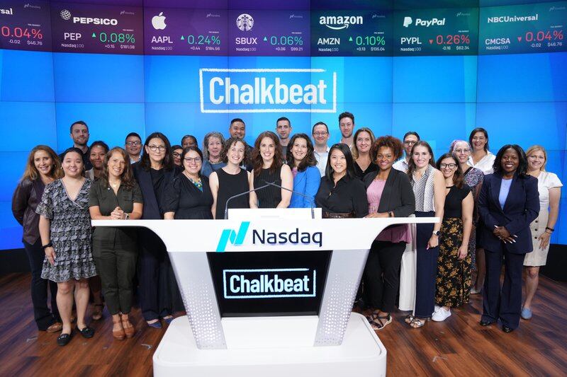 A group of about 30 people, mostly women, stands behind a lectern at Nasdaq. A blue screen with the Chalkbeat logo is in the background.