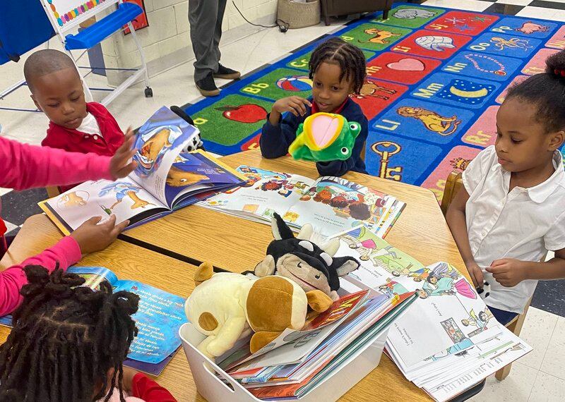 Three kindergarten read books together with their teacher at a table with books and hand puppets of different animals.